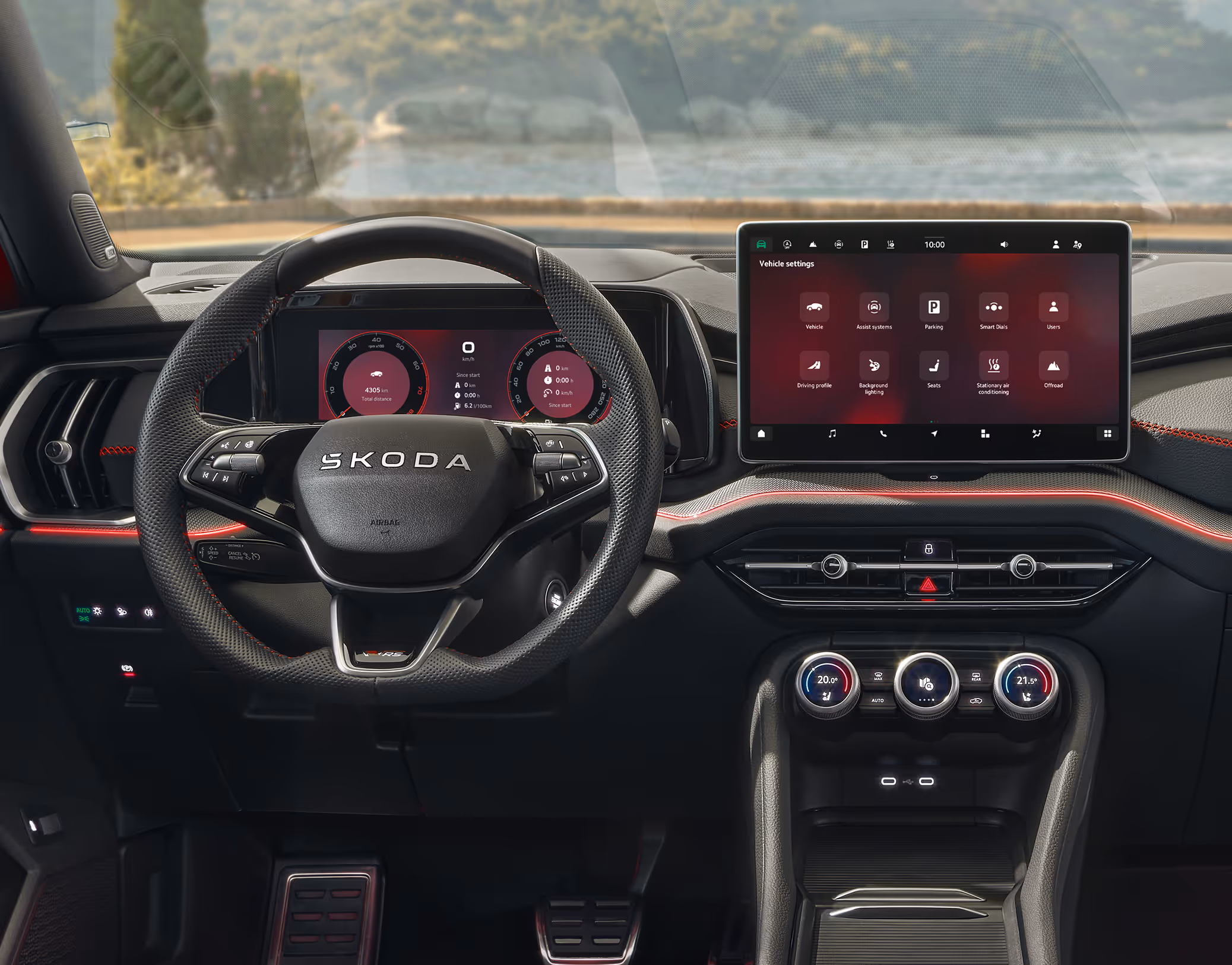 Eine frontale Ansicht des Cockpits eines Škoda Kodiaq RS. Das Sport-Multifunktionslenkrad, das digitale Cockpit und das große Infotainmentdisplay zeigen RS-spezifische rote Grafiken, die durch rote Kontrastnähte und eine Pedalerie in Edelstahloptik ergänzt werden. Durch die Windschutzscheibe ist eine malerische Küsten- und Berglandschaft zu sehen.