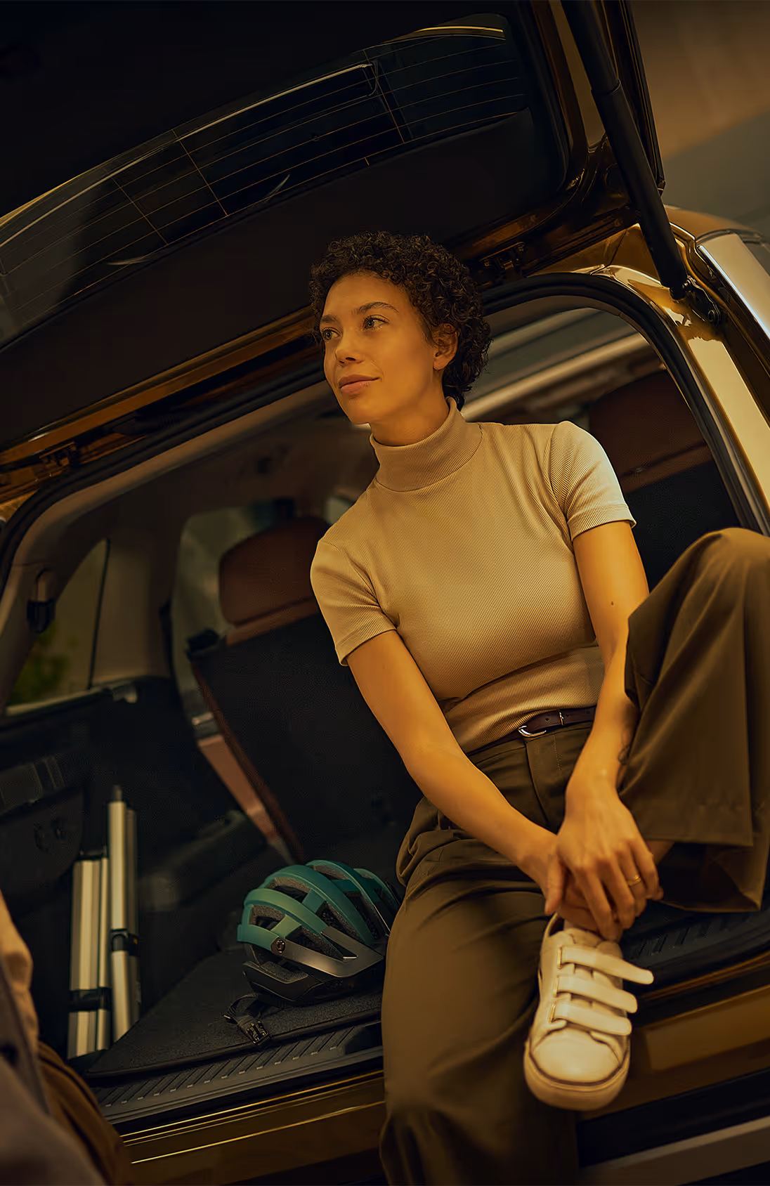 In einer modern beleuchteten Garage. Eine Frau sitzt entspannt auf der Ladekante des geöffneten Kofferraums, in dem ein Fahrradhelm liegt, während der Mann neben ihr steht. Der Škoda Kodiaq in der Lackierung Gold-Bronze Metallic hat die markanten Rückleuchten eingeschaltet. Unten rechts steht auf einer dunkelgrün abgesetzten Fläche "Individuelle Ausstattung" geschrieben.