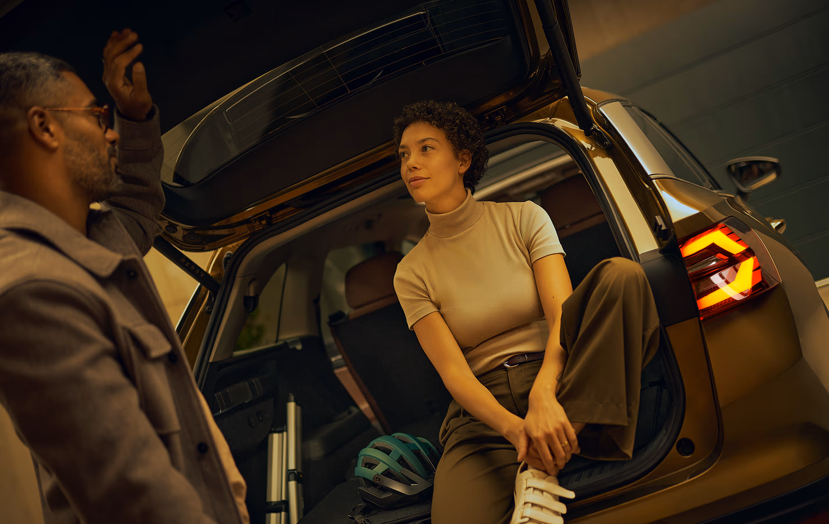 In einer modern beleuchteten Garage. Eine Frau sitzt entspannt auf der Ladekante des geöffneten Kofferraums, in dem ein Fahrradhelm liegt, während der Mann neben ihr steht. Der Škoda Kodiaq in der Lackierung Gold-Bronze Metallic hat die markanten Rückleuchten eingeschaltet. Unten rechts steht auf einer dunkelgrün abgesetzten Fläche "Individuelle Ausstattung" geschrieben.
