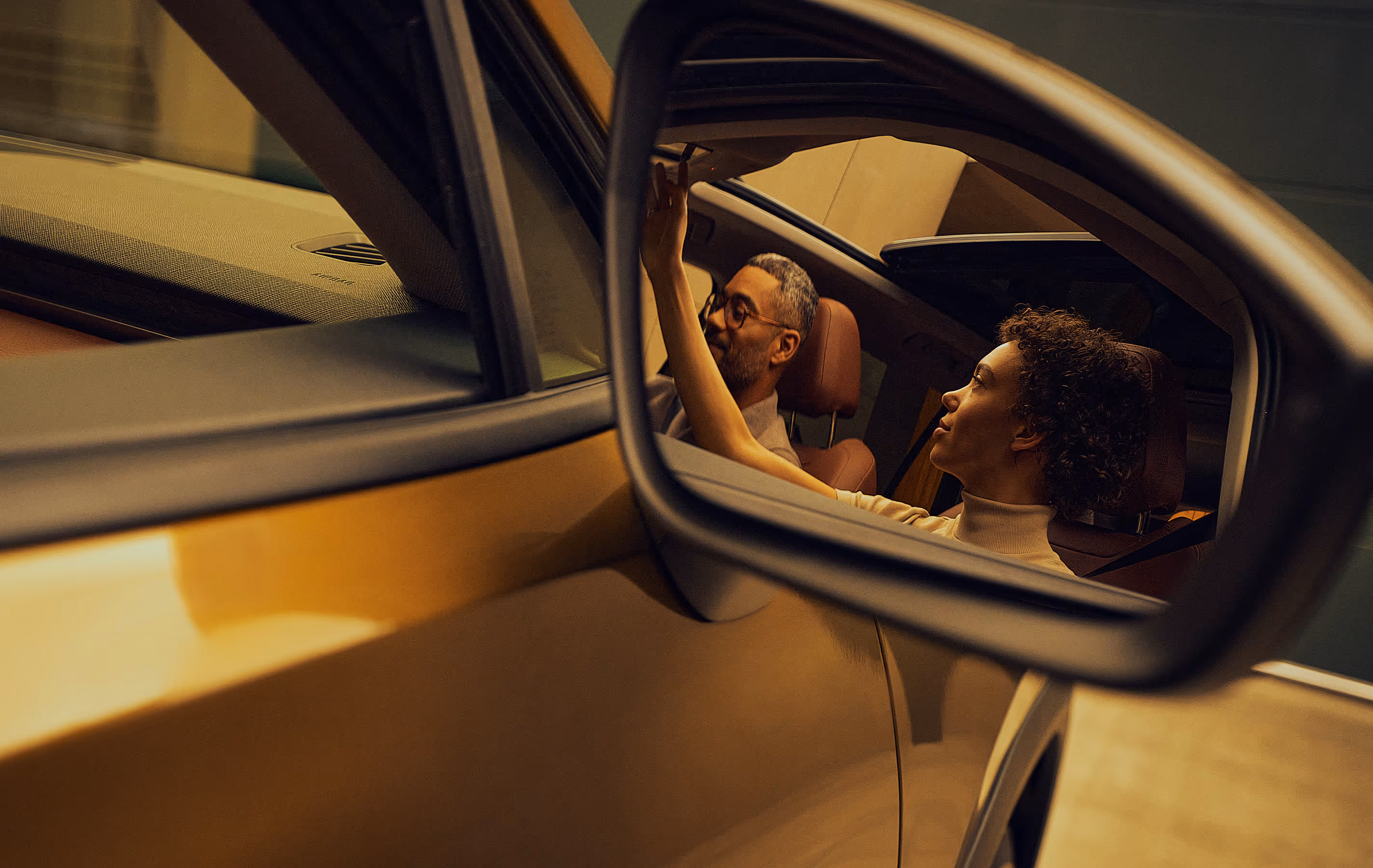 Eine Nahaufnahme des Außenspiegels eines Škoda Kodiaq in Gold-Bronze Metallic spiegelt eine Szene im Fahrzeuginneren wider. In der Spiegelung ist ein Paar zu sehen, das in den cognac-braunen Ledersitzen der Design Selection Lounge sitzt, während die Beifahrerin etwas am Dachhimmel einstellt. Unter rechts steht auf einer dunkelgrün abgesetzten Fläche geschrieben: "Sicherheit und Assistenten"