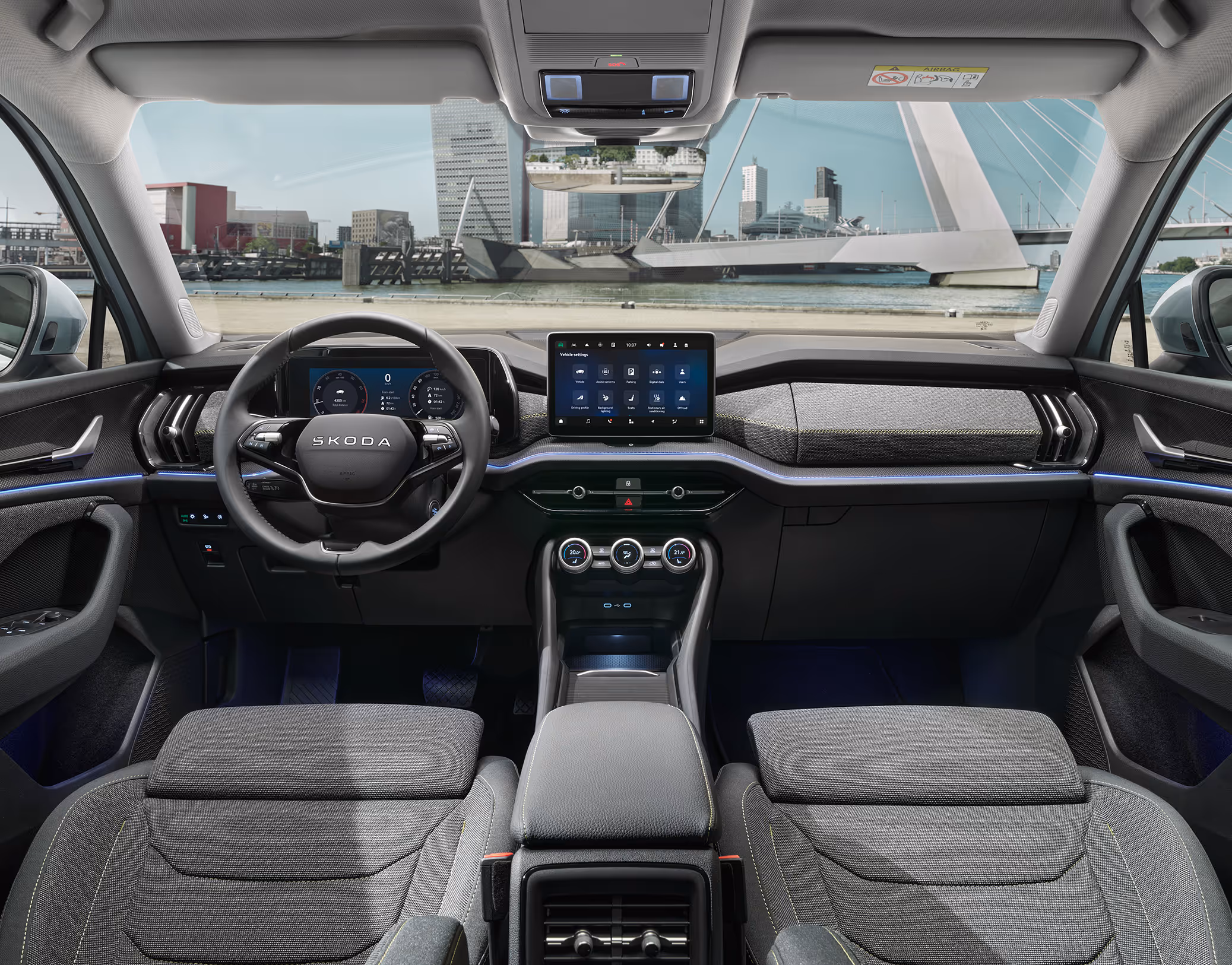 Eine weite Innenansicht aus der Rücksitzperspektive eines Škoda Kodiaq zeigt das gesamte Cockpit mit der Design Selection Loft. Das dunkle Interieur mit seinen grauen Stoffsitzen und einer Dekorleiste in Unique Dark Brushed steht im Kontrast zur hellen, städtischen Szenerie, die durch die Windschutzscheibe sichtbar ist.