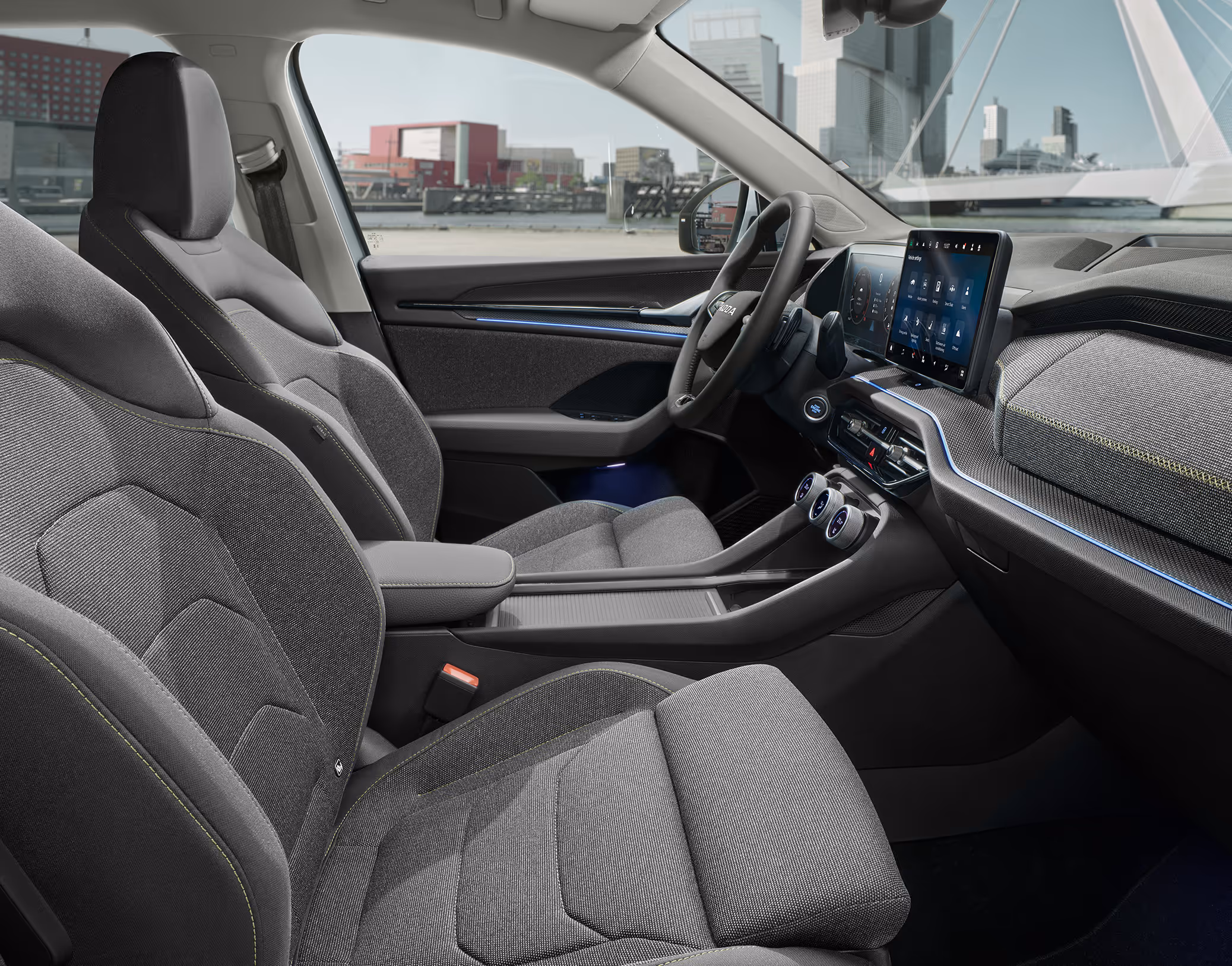 Eine seitliche Innenansicht des Škoda Kodiaq zeigt das Interieur der Design Selection Loft. Die grauen Stoffsitze sowie das dunkle Armaturenbrett mit einer Dekorleiste in Unique Dark Brushed sind zu sehen, während durch die Fenster eine helle, städtische Szenerie sichtbar ist.