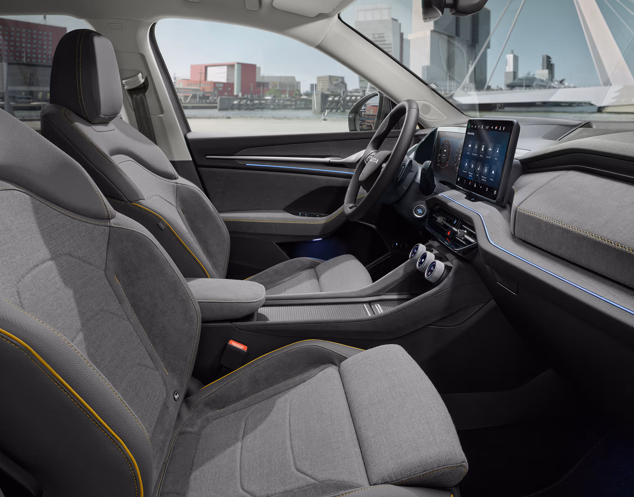Eine seitliche Innenansicht des Škoda Kodiaq zeigt das Interieur der Design Selection Lounge. Die Sitzbezüge aus einer grauen Suedia-Stoff-Kunstleder-Kombination mit gelben Kontrastnähten sowie das dunkle Armaturenbrett mit einer Dekorleiste in Unique Dark Brushed sind zu sehen, während durch die Fenster eine helle, städtische Szenerie sichtbar ist.