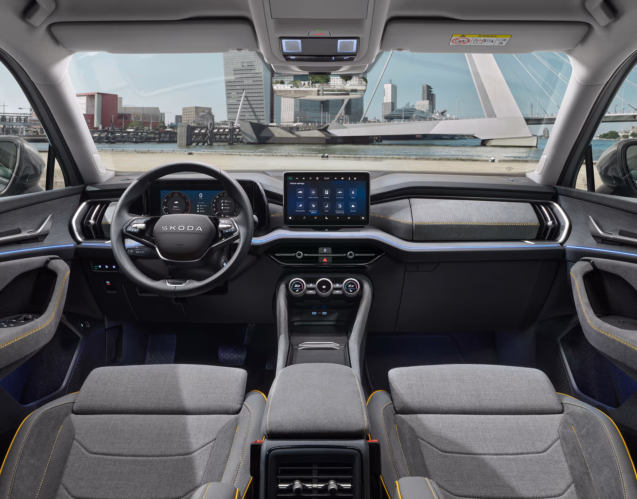 Eine weite Innenansicht aus der Rücksitzperspektive eines Škoda Kodiaq zeigt das gesamte Cockpit mit der Design Selection Lounge. Das dunkle Interieur mit seinen Sitzbezügen aus einer grauen Suedia-Stoff-Kunstleder-Kombination mit gelben Kontrastnähten und einer Dekorleiste in Unique Dark Brushed steht im Kontrast zur hellen, städtischen Szenerie, die durch die Windschutzscheibe sichtbar ist.
