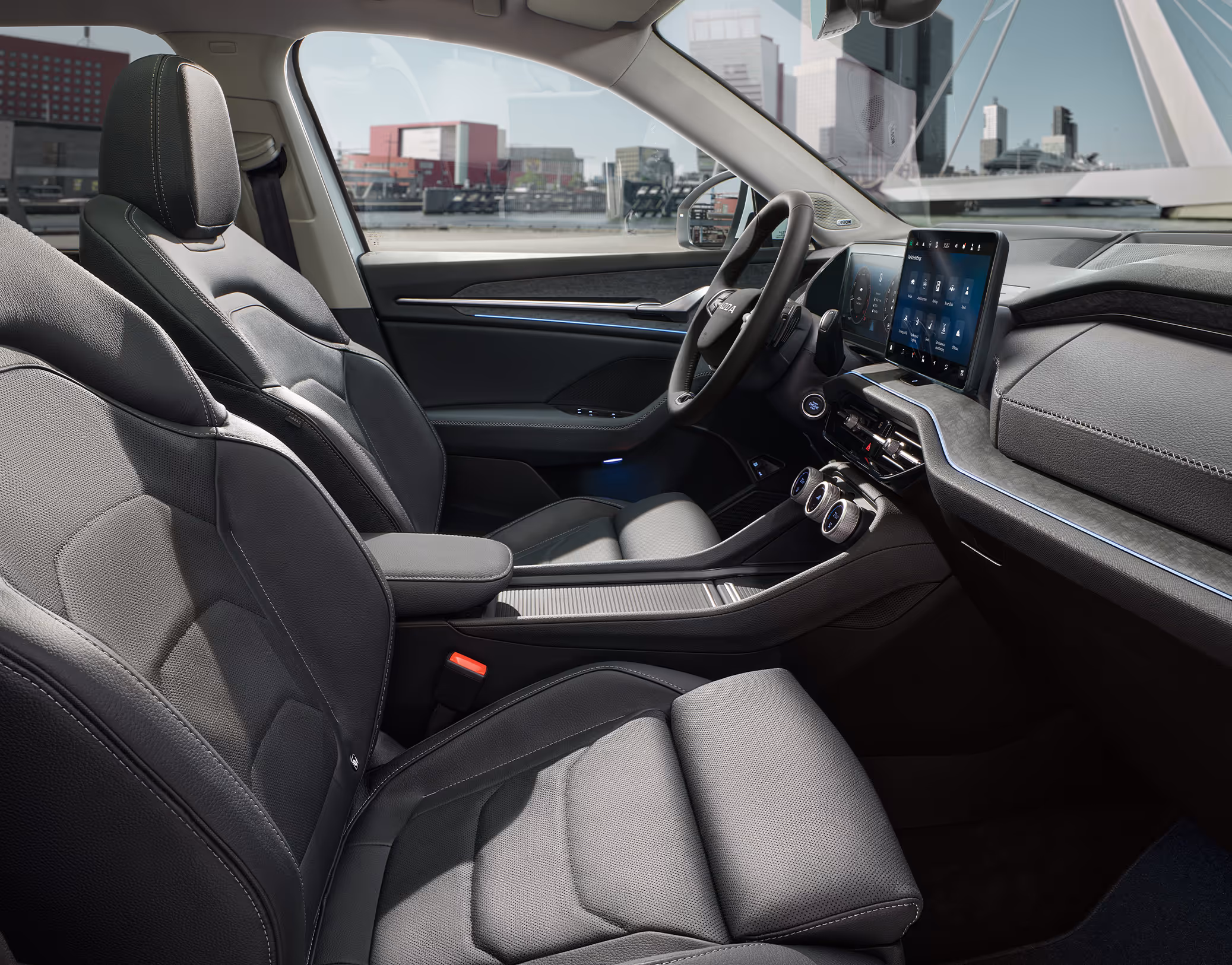 Eine seitliche Innenansicht des Škoda Kodiaq zeigt das Interieur der Design Selection Suite in Schwarz. Die schwarzen, perforierten Ledersitze mit grauen Kontrastnähten sowie das dunkle Armaturenbrett mit einer Dekorleiste in Unique Dark Brushed sind zu sehen, während durch die Fenster eine helle, städtische Szenerie sichtbar ist.