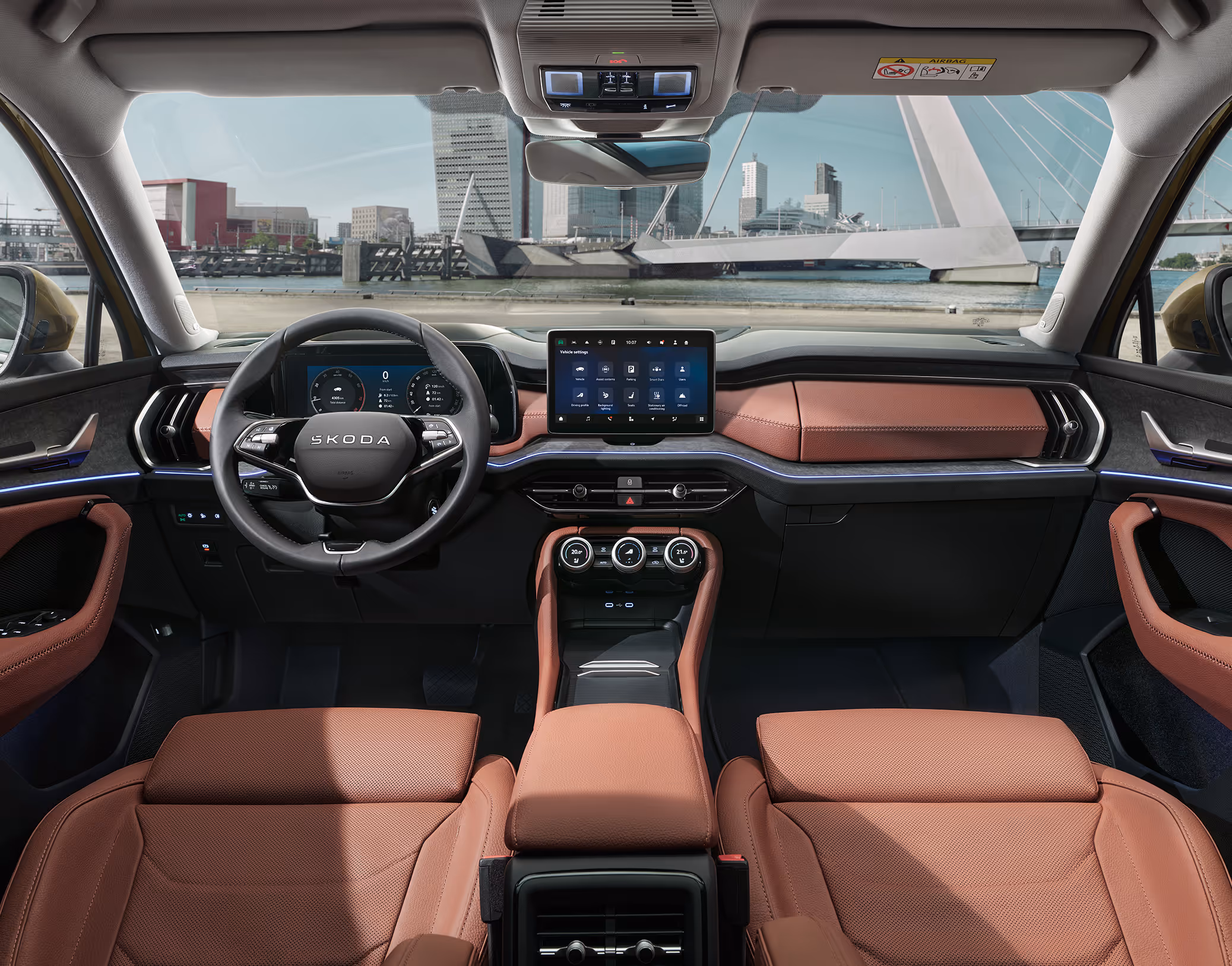 Eine weite Innenansicht aus der Rücksitzperspektive eines Škoda Kodiaq zeigt das gesamte Cockpit mit der Design Selection Suite in Cognac. Das Interieur mit seinen cognac-braunen Ledersitzen und einer Dekorleiste in Unique Dark Brushed steht im Kontrast zur hellen, städtischen Szenerie, die durch die Windschutzscheibe sichtbar ist.