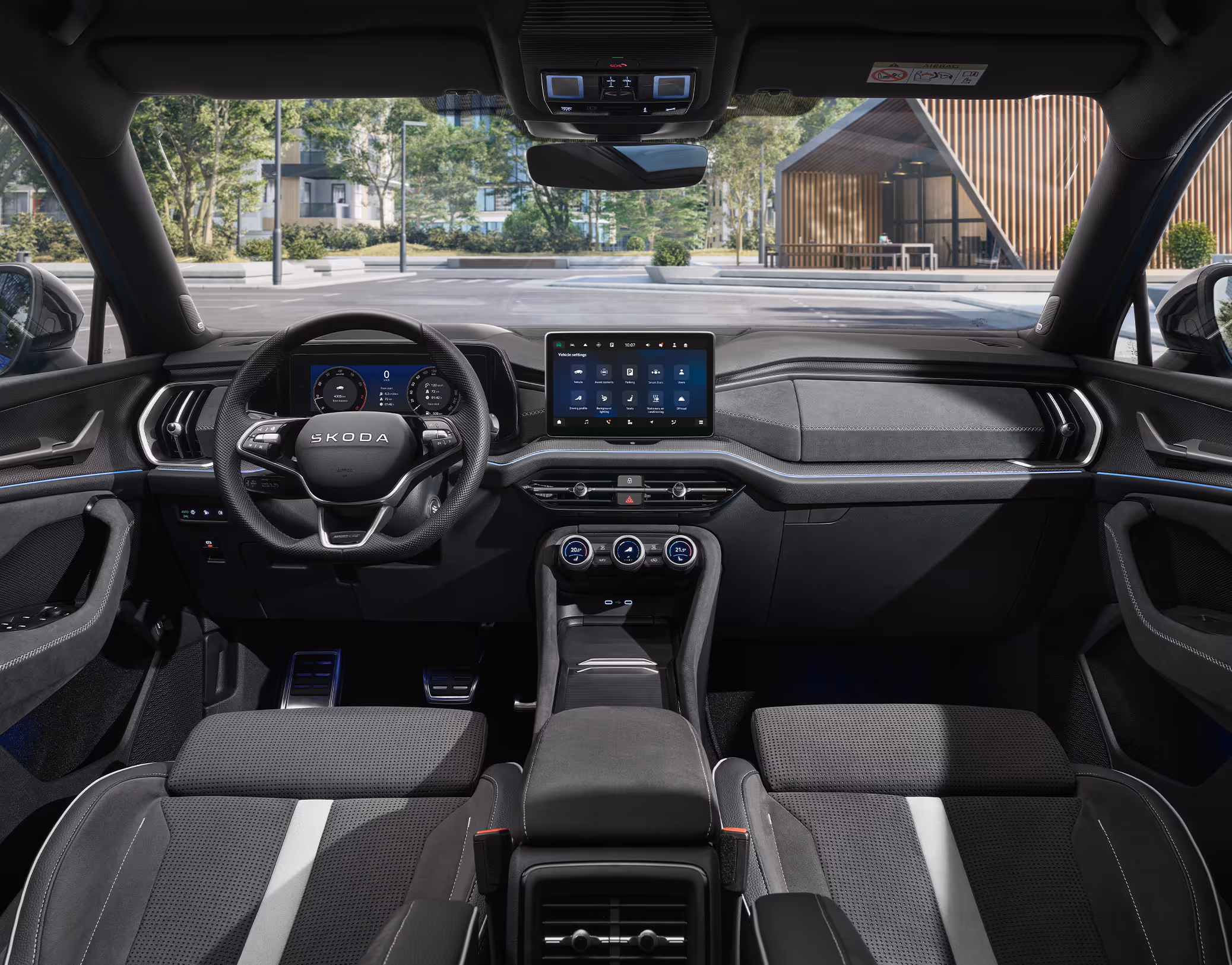 Eine weite Innenansicht aus der Rücksitzperspektive eines Škoda Kodiaq Sportline zeigt das gesamte Cockpit und die vorderen Sportsitze. Das dunkle Interieur der Design Selection Sportline mit seinen markanten Sitzen, dem digitalen Cockpit und dem großen Infotainmentdisplay steht im Kontrast zur hellen, städtischen Szenerie, die durch die Windschutzscheibe sichtbar ist.