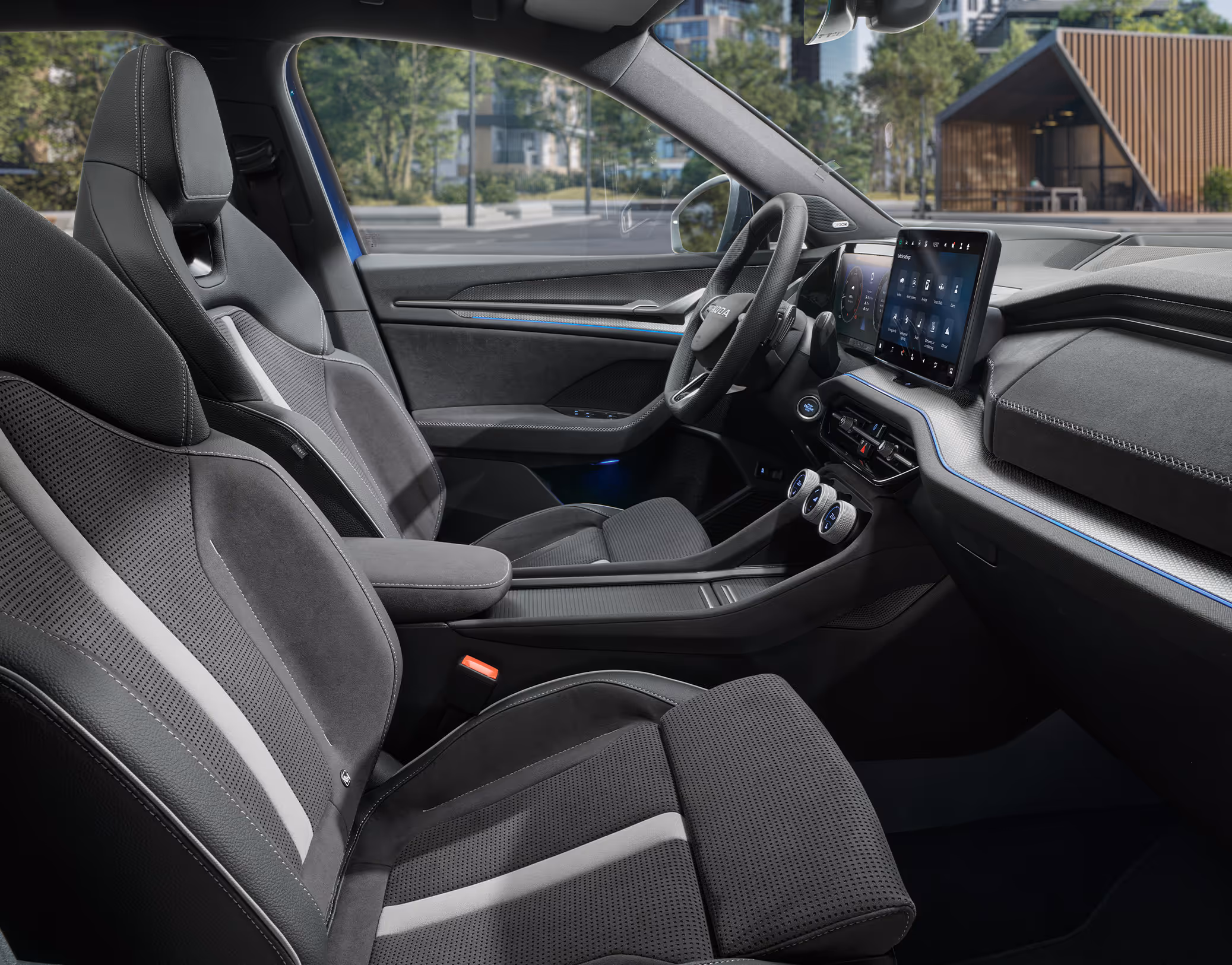 Eine seitliche Innenansicht des Škoda Kodiaq Sportline zeigt das sportliche Interieur der Design Selection Sportline. Die schwarzen Sportsitze mit integrierten Kopfstützen, weiß-gräulichen Zierstreifen und Kontrastnähten prägen den dynamischen Charakter des Cockpits. Durch die Fenster ist eine helle, städtische Szenerie zu sehen, die einen Kontrast zum dunklen Innenraum bildet.