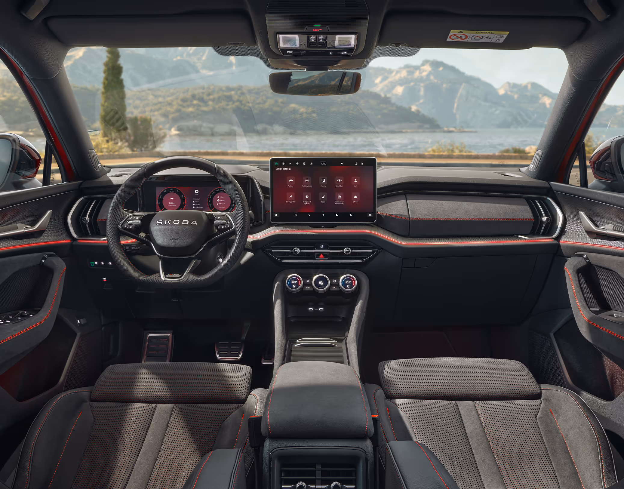 Eine weite Innenansicht aus der Rücksitzperspektive eines Škoda Kodiaq zeigt das gesamte Cockpit mit der Design Selection RS Lounge. Das sportliche, dunkle Interieur mit seinen Sitzbezügen aus schwarzem Suedia-Stoff mit roten Kontrastnähten und Dekorleisten in Carbon-Optik steht im Kontrast zur hellen, städtischen Szenerie, die durch die Windschutzscheibe sichtbar ist.