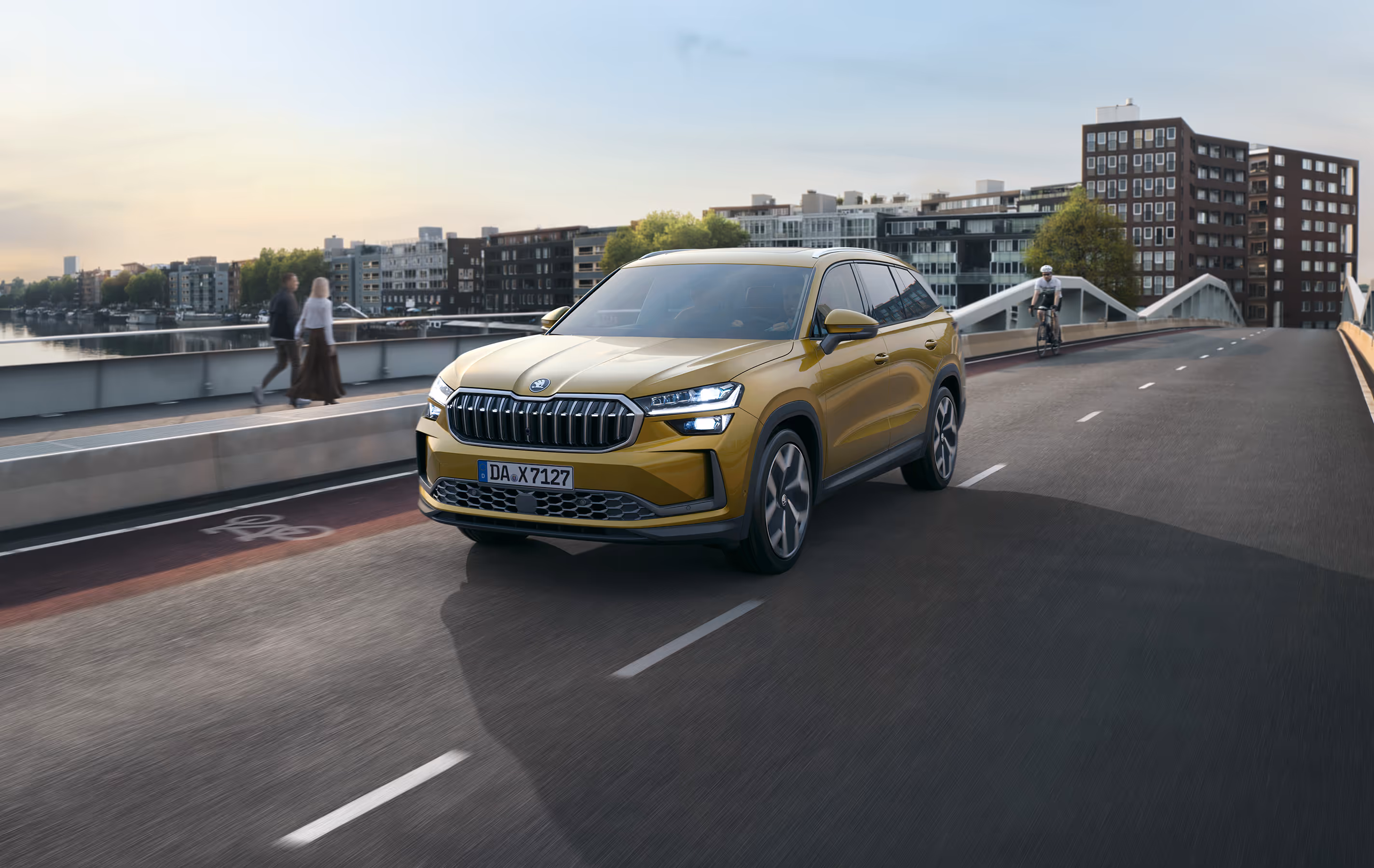 Ein Škoda Kodiaq in der Lackierung Gold-Bronze Metallic fährt auf einer Brücke durch eine städtische Umgebung. Die Szene ist in das warme Licht der tiefstehenden Sonne getaucht, und im Hintergrund sind moderne Gebäude und ein Wasserweg zu sehen. Oben links im Bild steht weiß "SKODA" geschrieben, unten rechts auf einer dunkelgrün abgesetzten Fläche steht "Kodiaq".