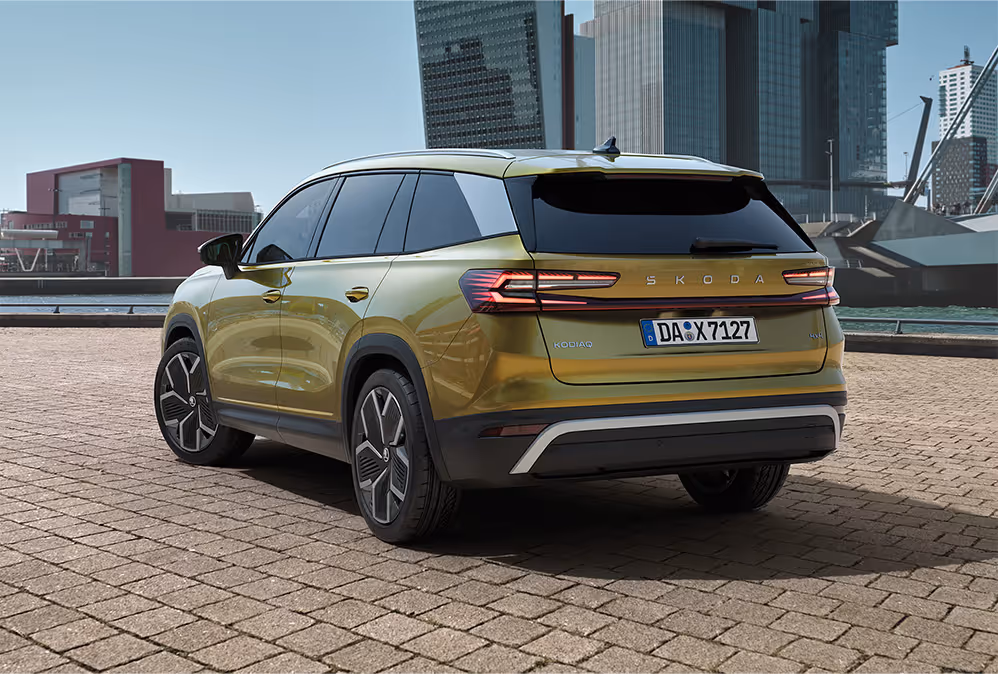 Ein Škoda Kodiaq in der Lackierung Gold-Bronze Metallic ist von schräg hinten auf einem gepflasterten Platz zu sehen. Das Heckdesign wird durch die durchgehende rote Leiste zwischen den LED-Heckleuchten und den neuen Škoda Schriftzug geprägt. Im Hintergrund erhebt sich die beeindruckende Skyline einer modernen Hafenstadt.