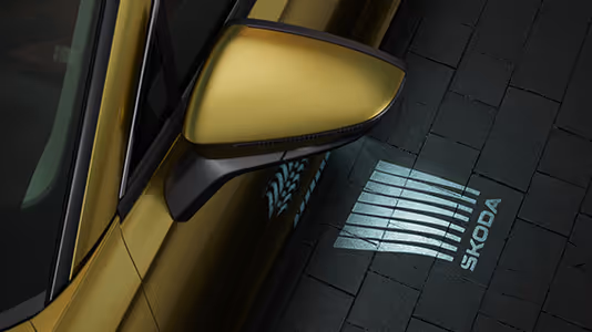 Eine Nahaufnahme zeigt den Außenspiegel eines Škoda Kodiaq in der Lackierung Gold-Bronze Metallic bei Nacht. Aus dem Spiegel wird das Welcome-Logo, eine Grafik mit dem Škoda Schriftzug, auf den dunklen, gepflasterten Boden projiziert. Diese Einstiegsbeleuchtung erhellt den Bereich neben der Tür und begrüßt die Passagiere.