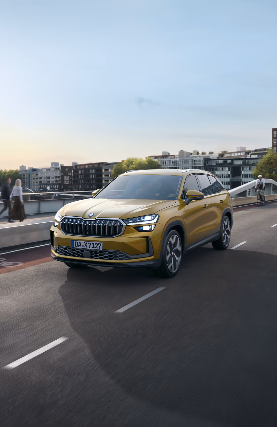 Ein Škoda Kodiaq in der Lackierung Gold-Bronze Metallic fährt auf einer Brücke durch eine städtische Umgebung. Die Szene ist in das warme Licht der tiefstehenden Sonne getaucht, und im Hintergrund sind moderne Gebäude und ein Wasserweg zu sehen. Oben links im Bild steht weiß "SKODA" geschrieben, unten rechts auf einer dunkelgrün abgesetzten Fläche steht "Kodiaq".