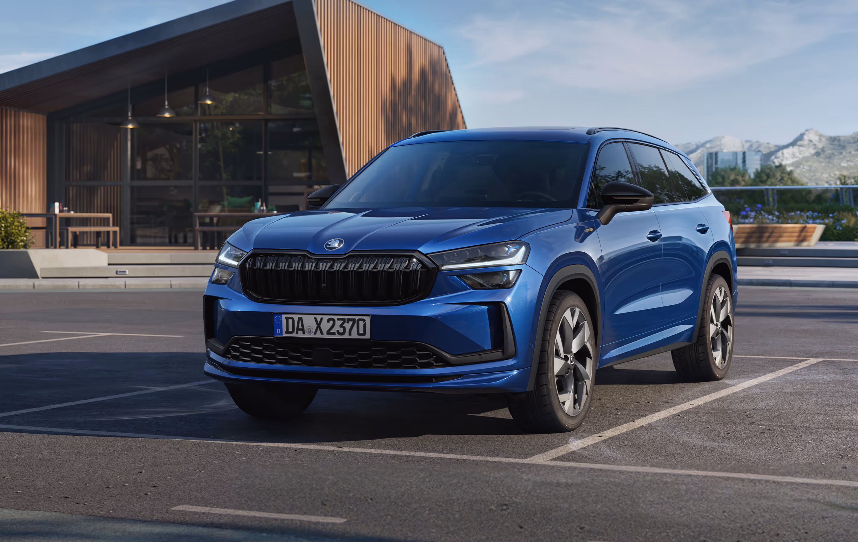 Ein Škoda Kodiaq in der Ausstattungslinie Sportline und der Lackierung Race-Blau Metallic parkt auf einem Parkplatz vor einem modernen Gebäude. Im Hintergrund der sonnigen Szene sind eine Berglandschaft und Bäume zu erkennen. Rechts unten steht auf einer dunkelgrün abgesetzten Fläche "Sportline" geschrieben.