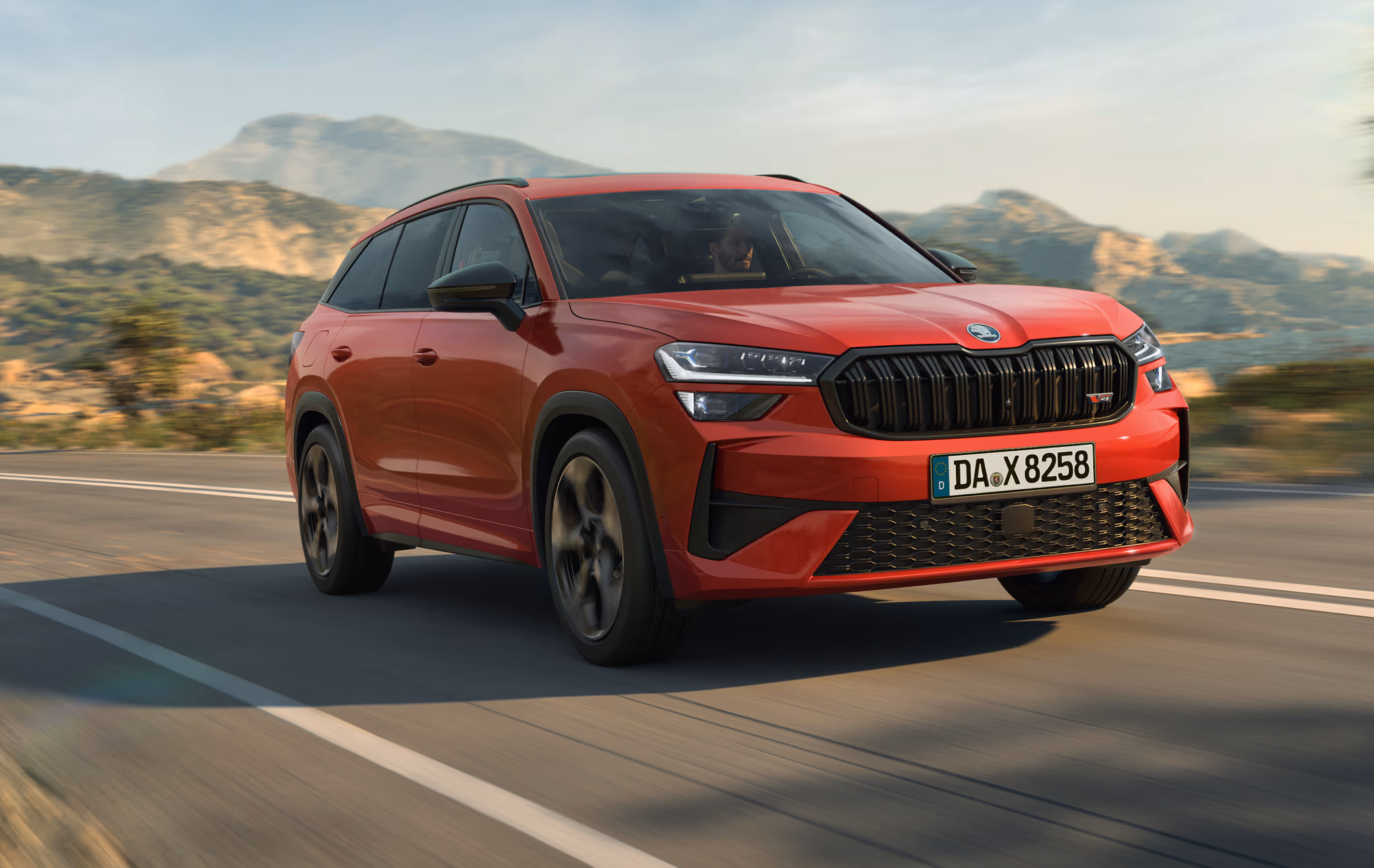 Ein Škoda Kodiaq RS in der Lackierung Velvet-Rot Metallic fährt auf einer kurvenreichen Straße durch eine hügelige Landschaft. Die Aufnahme bei tiefstehender Sonne zeigt das Fahrzeug in einer Dreiviertelansicht von vorne und betont die sportlichen Designelemente wie den schwarzen Kühlergrill mit RS-Logo. Der durch Bewegungsunschärfe dargestellte Hintergrund unterstreicht den dynamischen Charakter der Szene.