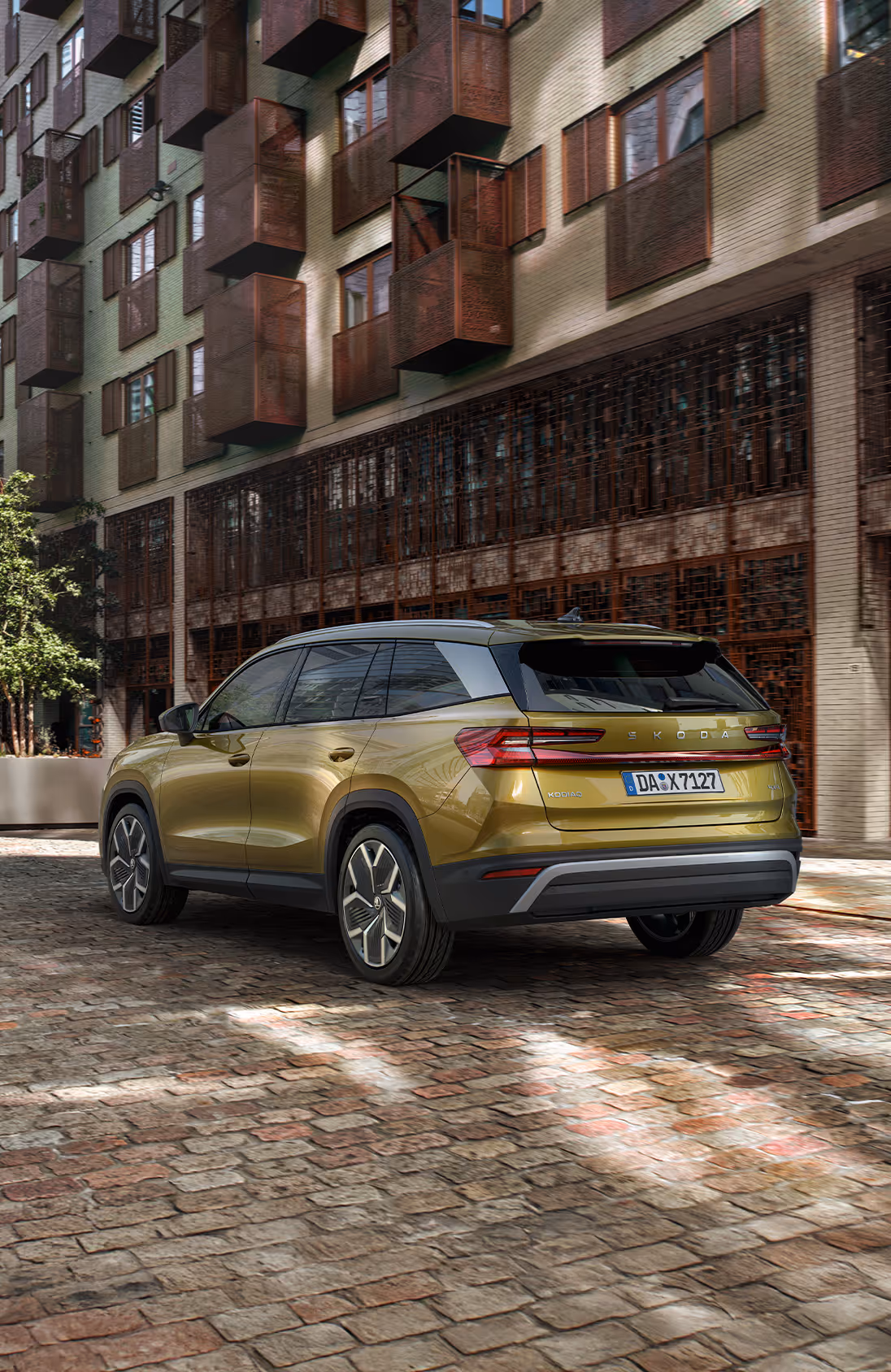 Ein Škoda Kodiaq in der Lackierung Gold-Bronze Metallic ist auf einer Kopfsteinpflasterstraße in einer städtischen Umgebung geparkt. Das Fahrzeug wird von schräg hinten gezeigt, vor der Fassade eines modernen Gebäudes, während eine Frau auf dem Gehweg daran vorbeigeht. Rechts unten im Bild steht auf einer dunkelgrün abgesetzten Fläche "Exterieur-Design" geschrieben.