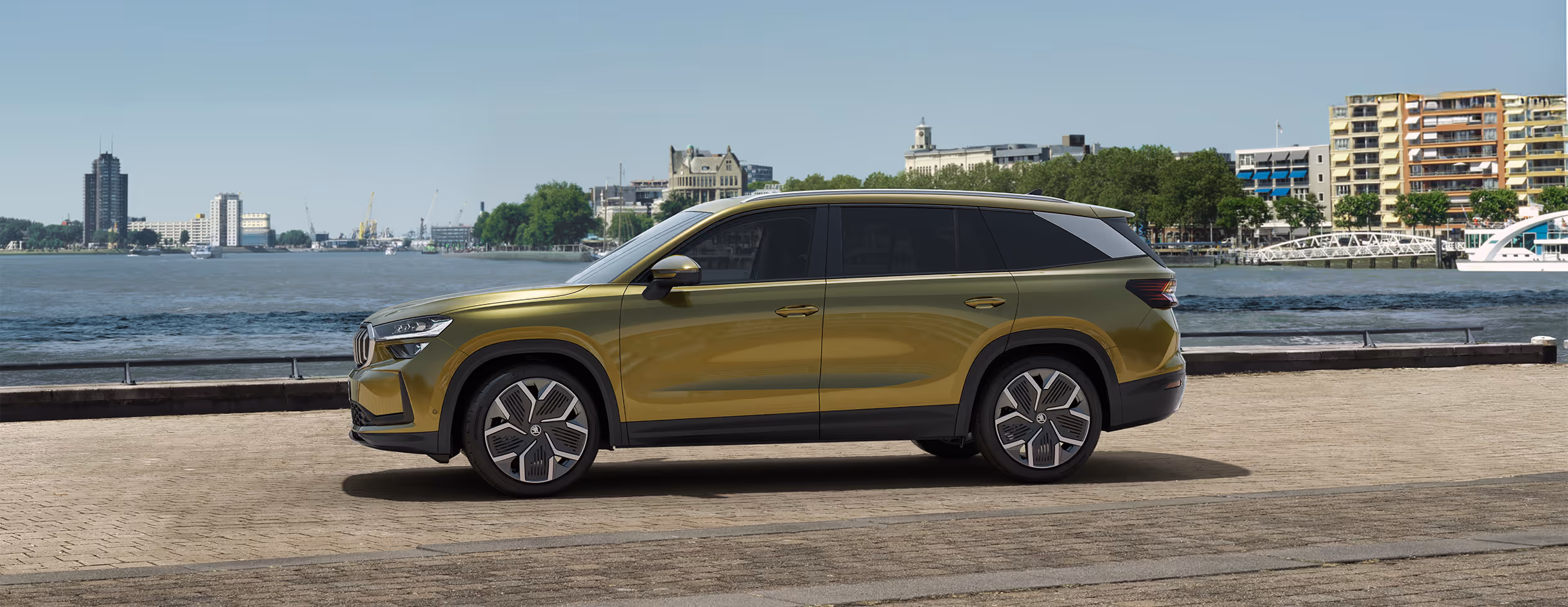 Ein Škoda Kodiaq in der Lackierung Gold-Bronze Metallic ist in der Seitenansicht an einer Uferpromenade abgebildet. Im Hintergrund sind ein Gewässer und die Silhouette einer Stadt mit Hafenanlagen zu erkennen. Die gestreckte Seitenlinie des SUVs wird durch die dunklen Fenster, das dunkle Dach und die markanten Leichtmetallfelgen betont.