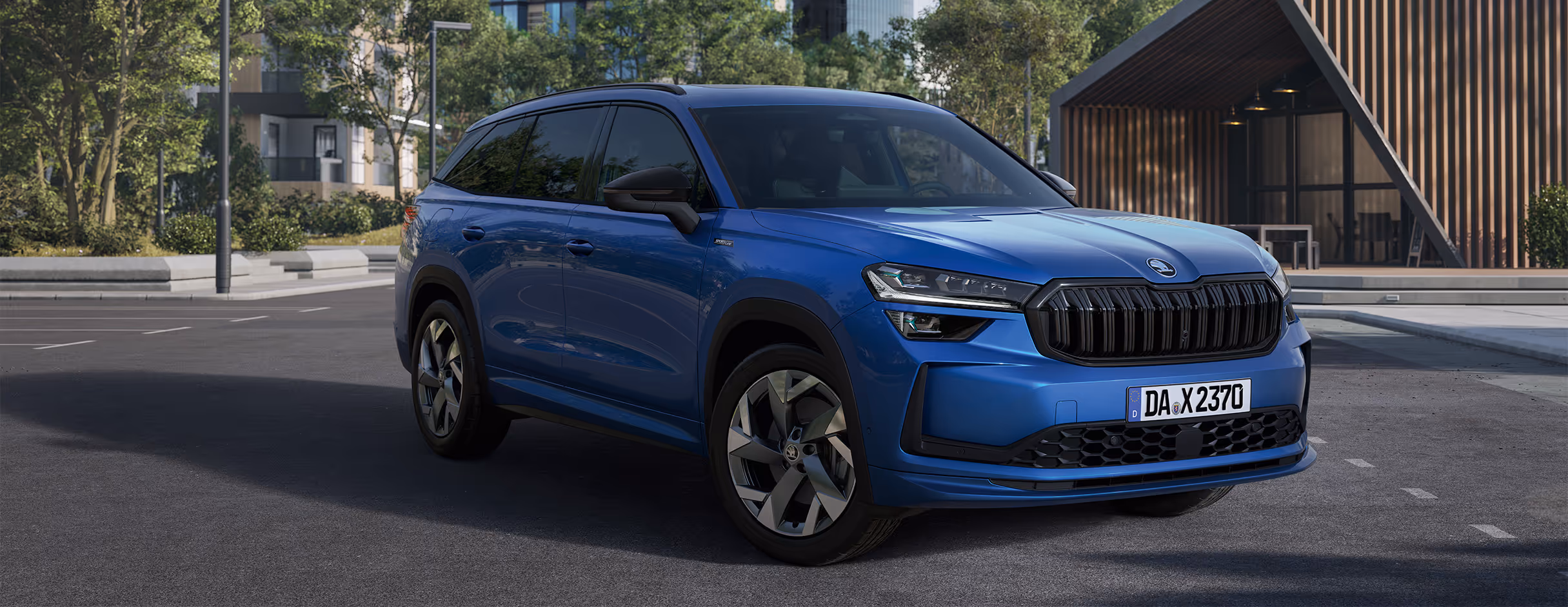 Eine Nahaufnahme zeigt die Frontpartie eines Škoda Kodiaq Sportline in der Lackierung Race-Blau Metallic. Die sportliche Front wird durch den schwarzen Škoda Kühlergrill und die scharf geschnittenen TOP LED-Matrix-Scheinwerfer betont. Das Fahrzeug parkt vor einer modernen Architektur mit Holzelementen, während im Hintergrund Bäume und städtische Gebäude zu sehen sind.