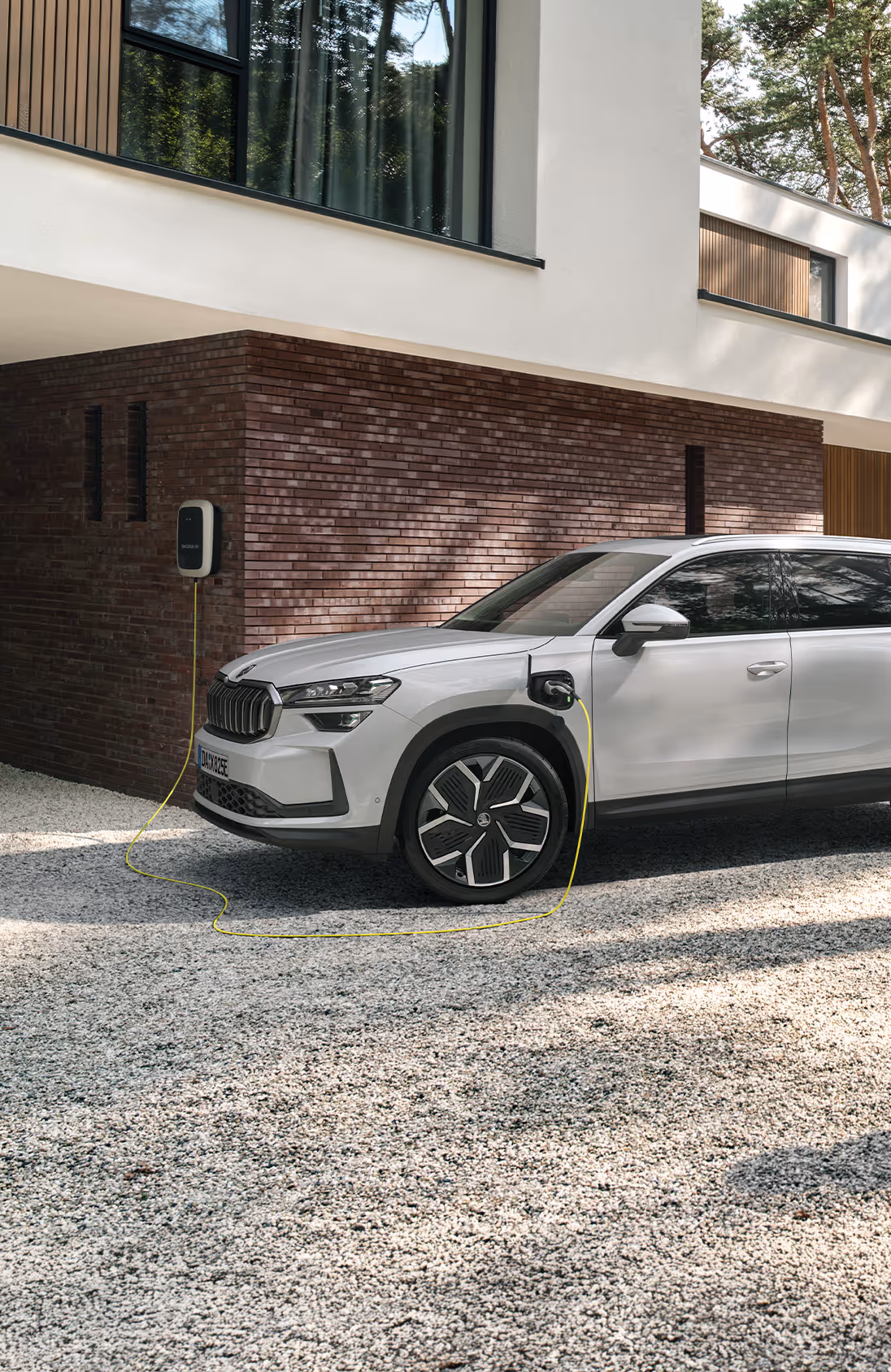 Ein Škoda Kodiaq iV Plug-in-Hybrid mit der Lackierung Moon-Weiß Perleffekt lädt an einer Wallbox in der Kieseinfahrt eines modernen Hauses. Das Fahrzeug ist über ein Ladekabel mit der Ladestation verbunden, während im Vordergrund ein kleines Kind auf einen Ball zuläuft. Rechts unten steht auf einer dunkelgrün abgesetzten Fläche "Kodiaq iV" geschrieben.
