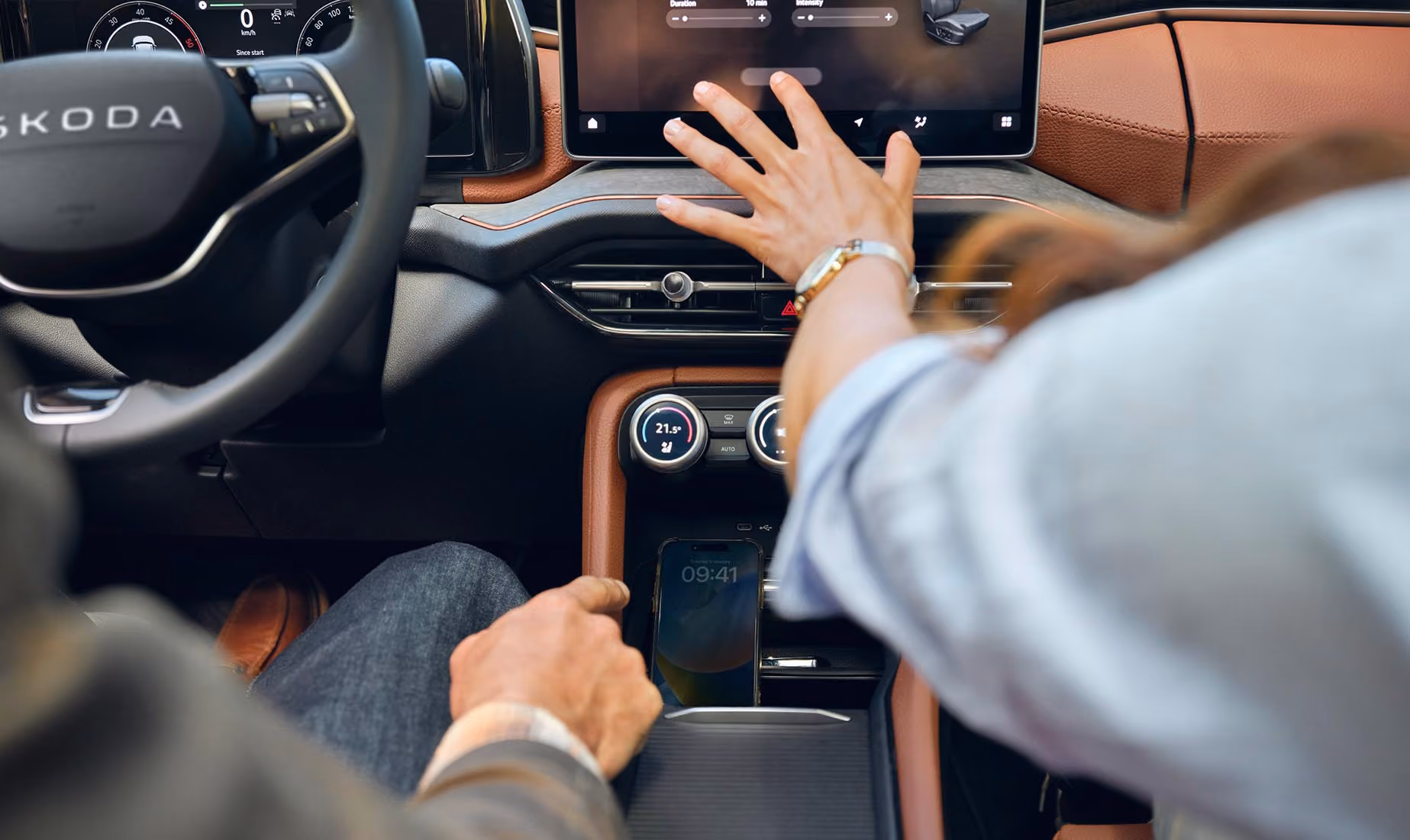 Blick von der Rückbank aus auf ein Paar im vorderen Teil eines Škoda Kodiaq. Die Beifahrerin interagiert mit dem großen Infotainmentdisplay und zeigt auf die Navigationskarte, während der Fahrer ebenfalls dorthin schaut. Das Interieur mit den cognac-braunen Ledersitzen der Design Selection Lounge ist stimmungsvoll beleuchtet.  Unten rechts steht auf einer dunkelgrün abgesetzten Fläche geschrieben: "Škoda Connect"