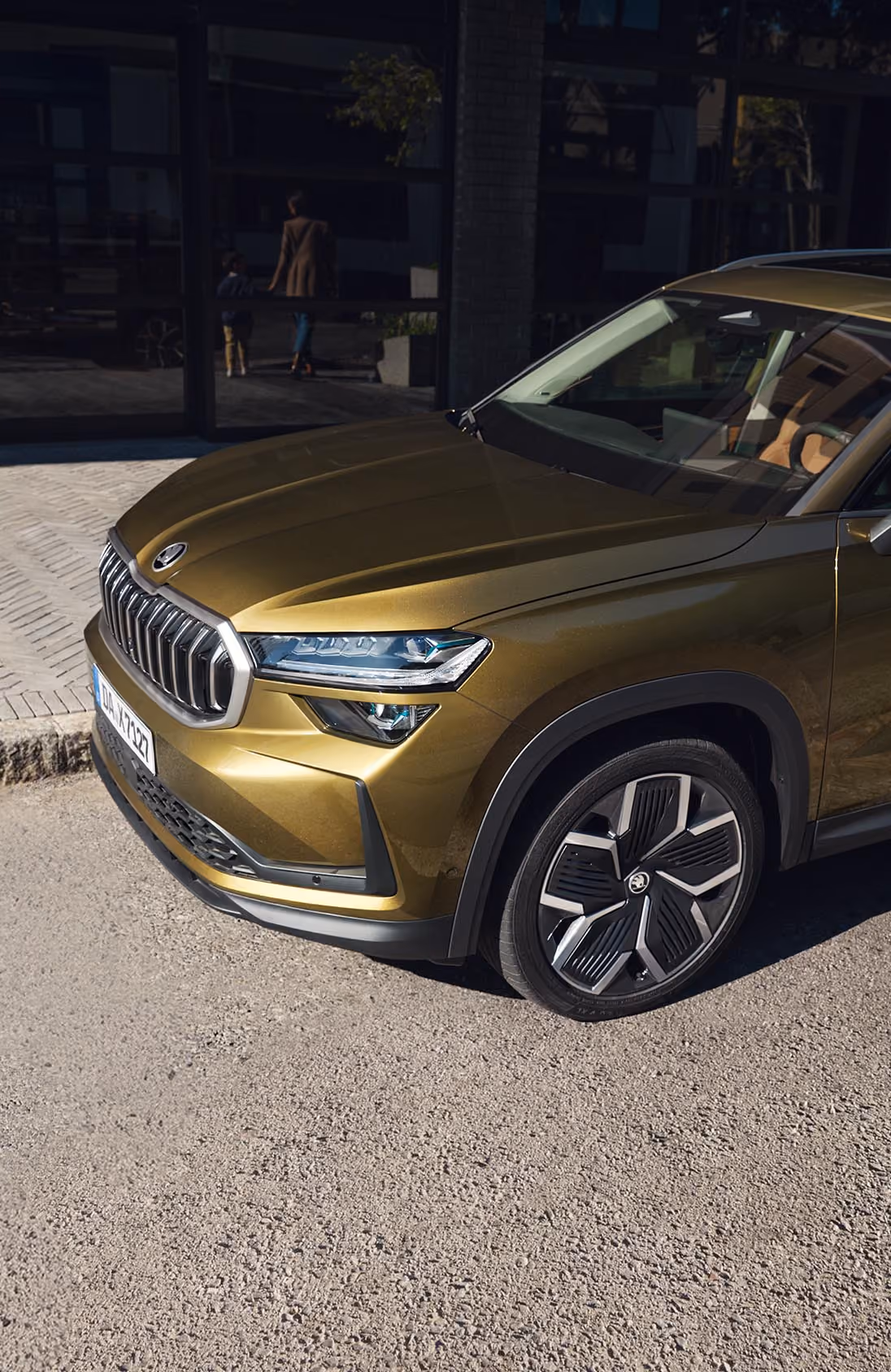 Blick aus einer erhöhten Perspektive auf einen Škoda Kodiaq in der Lackierung Gold-Bronze Metallic, der in einer modernen Garage parkt. Eine Frau steigt auf der Beifahrerseite ein, während ein Mann gleichzeitig die Fahrertür öffnet. An der Garagenwand ist ein sportliches Škoda Fahrrad mit silbernem Rahmen befestigt. Die Deckenbeleuchtung spiegelt sich auf dem Fahrzeugdach, der Windschutzscheibe und der Motorhaube.