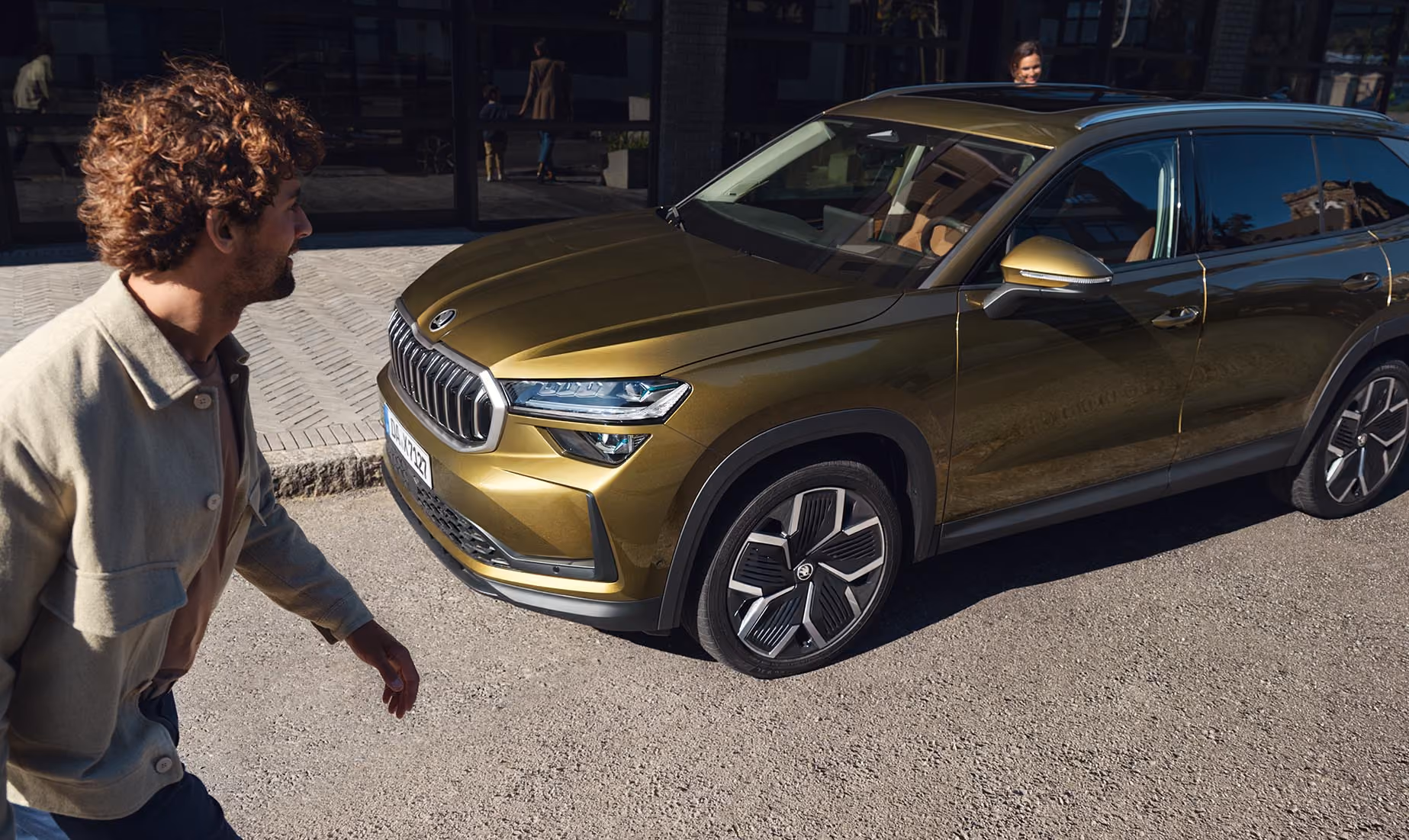 Blick aus einer erhöhten Perspektive auf einen Škoda Kodiaq in der Lackierung Gold-Bronze Metallic, der in einer modernen Garage parkt. Eine Frau steigt auf der Beifahrerseite ein, während ein Mann gleichzeitig die Fahrertür öffnet. An der Garagenwand ist ein sportliches Škoda Fahrrad mit silbernem Rahmen befestigt. Die Deckenbeleuchtung spiegelt sich auf dem Fahrzeugdach, der Windschutzscheibe und der Motorhaube.
