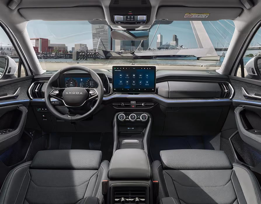 Eine weite Innenansicht aus der Rücksitzperspektive eines Škoda Kodiaq zeigt das gesamte Cockpit mit der Design Selection Suite in Schwarz. Das dunkle Interieur mit seinen schwarzen Ledersitzen und einer Dekorleiste in Unique Dark Brushed steht im Kontrast zur hellen, städtischen Szenerie, die durch die Windschutzscheibe sichtbar ist.