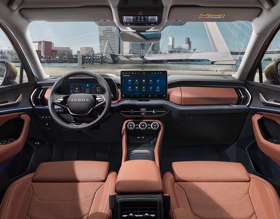 Eine weite Innenansicht aus der Rücksitzperspektive eines Škoda Kodiaq zeigt das gesamte Cockpit mit der Design Selection Suite in Cognac. Das Interieur mit seinen cognac-braunen Ledersitzen und einer Dekorleiste in Unique Dark Brushed steht im Kontrast zur hellen, städtischen Szenerie, die durch die Windschutzscheibe sichtbar ist.