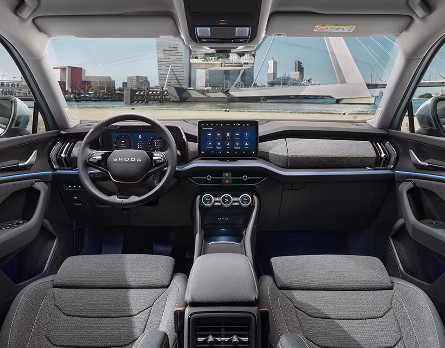 Eine weite Innenansicht aus der Rücksitzperspektive eines Škoda Kodiaq zeigt das gesamte Cockpit mit der Design Selection Loft. Das dunkle Interieur mit seinen grauen Stoffsitzen und einer Dekorleiste in Unique Dark Brushed steht im Kontrast zur hellen, städtischen Szenerie, die durch die Windschutzscheibe sichtbar ist.