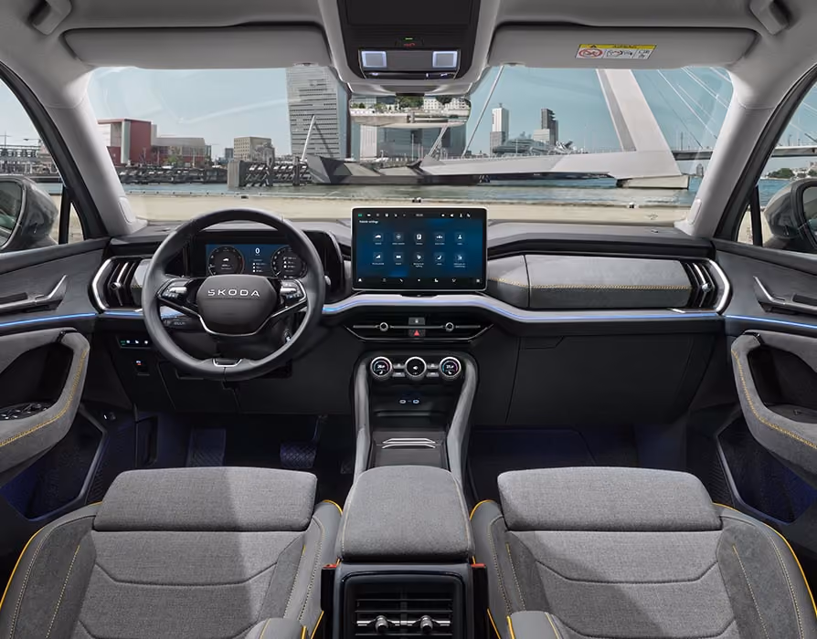 Eine weite Innenansicht aus der Rücksitzperspektive eines Škoda Kodiaq zeigt das gesamte Cockpit mit der Design Selection Lounge. Das dunkle Interieur mit seinen Sitzbezügen aus einer grauen Suedia-Stoff-Kunstleder-Kombination mit gelben Kontrastnähten und einer Dekorleiste in Unique Dark Brushed steht im Kontrast zur hellen, städtischen Szenerie, die durch die Windschutzscheibe sichtbar ist.