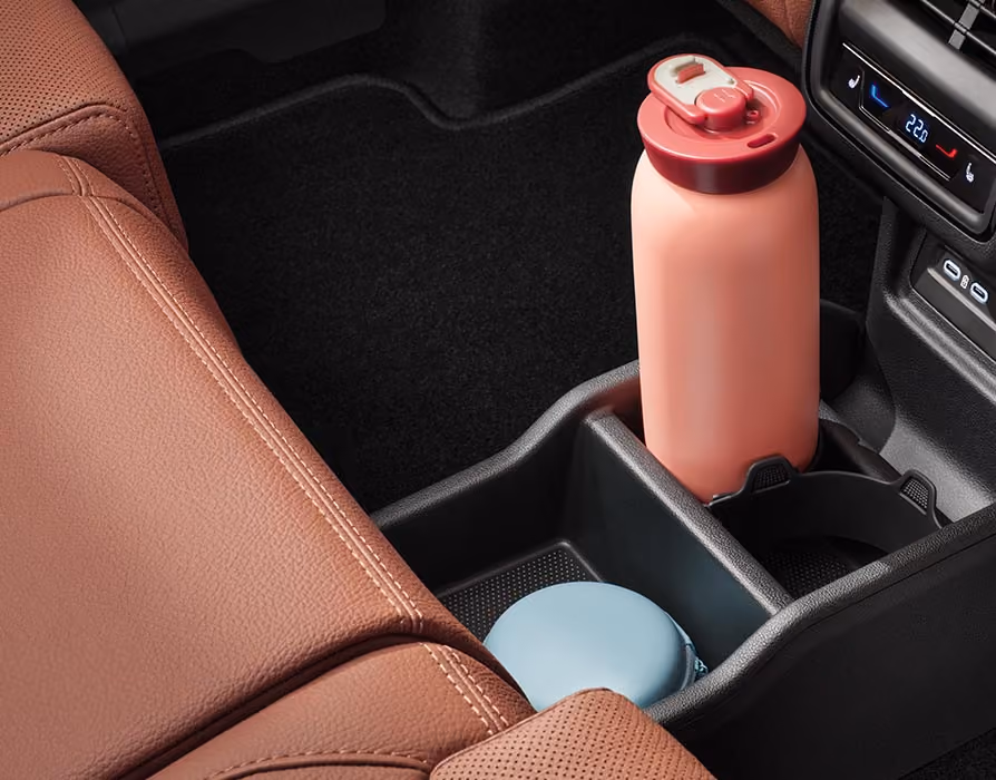 Eine Nahaufnahme zeigt den hinteren Teil der Mittelkonsole im Škoda Kodiaq. Unter den Lüftungsdüsen und den Bedienelementen für die 3-Zonen-Klimaanlage befinden sich zwei USB-C-Anschlüsse und ein großes Ablagefach mit Getränkehalter. Die cognac-braunen Ledersitze der Design Selection Lounge rahmen die Szene ein.