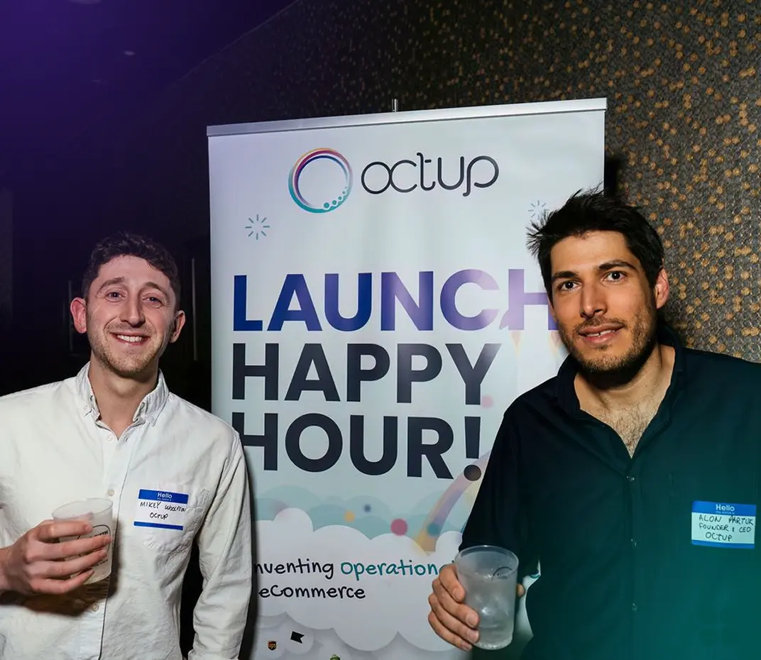 Two men holding plastic cups stand in front of a banner reading 'Octup Launch Happy Hour!' at an event.