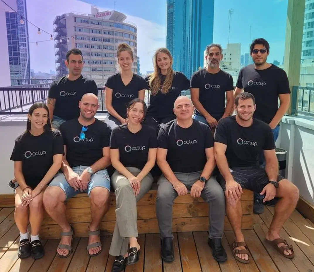 Group of ten people wearing black Octup t-shirts sitting and standing on a balcony with city buildings in the background.