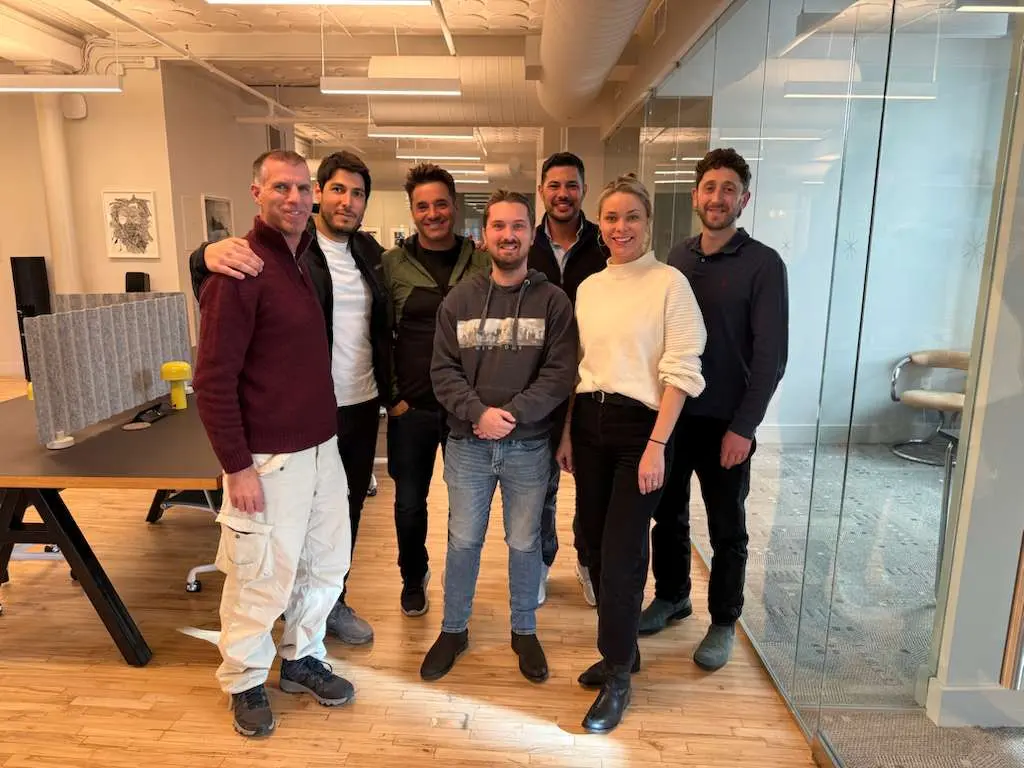 A group of seven diverse people standing closely together in a modern office with wooden floors and glass walls, smiling at the camera.