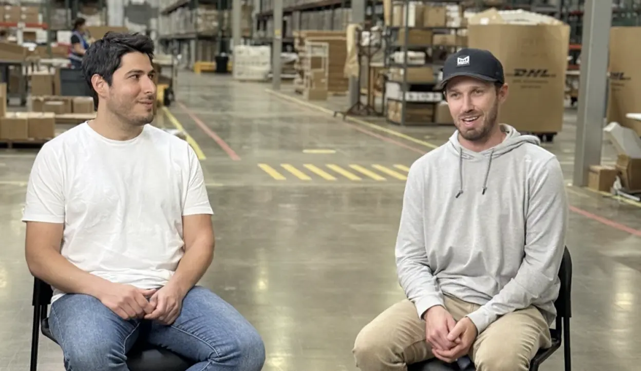 Two men sitting and talking in a warehouse with shelves and boxes in the background.