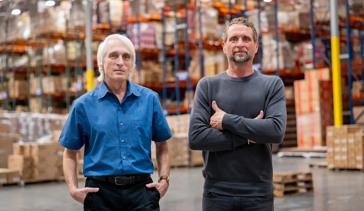 Two men standing in a warehouse with shelves stocked with boxes; one man wears a blue shirt with hands in pockets, and the other wears a gray long-sleeve shirt with arms crossed.