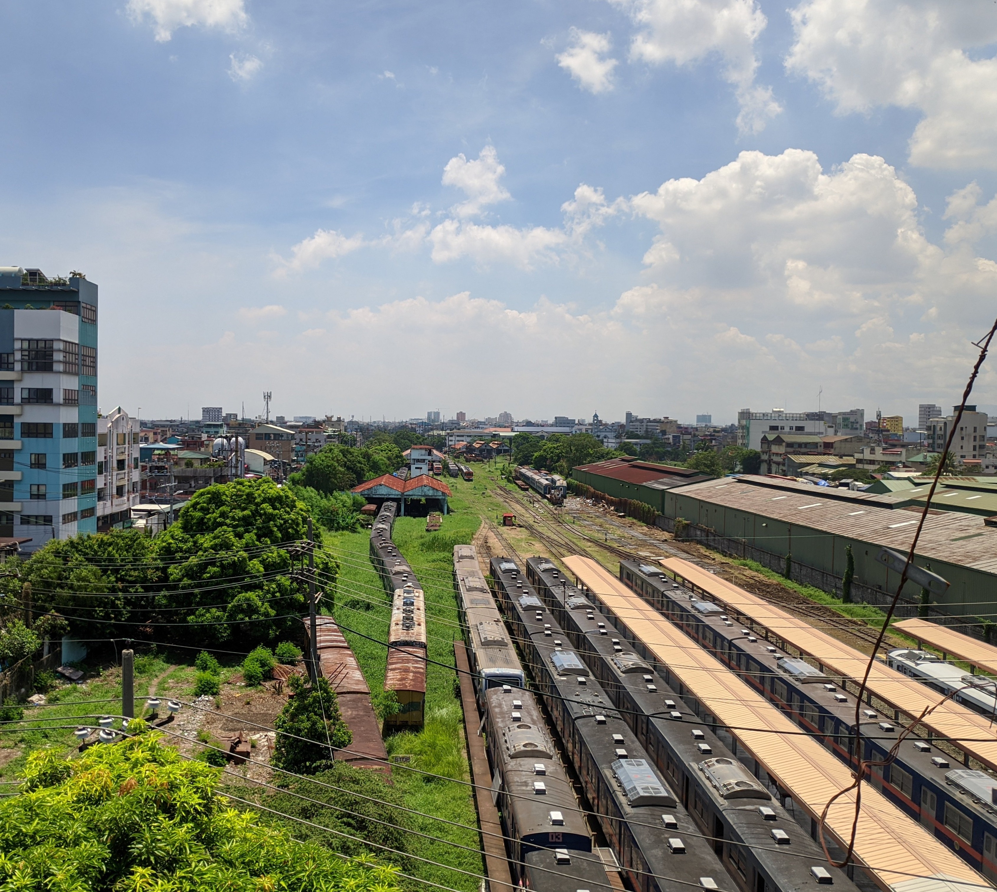 Aerial view of multiple train cars parked on railway tracks with green trees and buildings in the background under a partly cloudy sky.