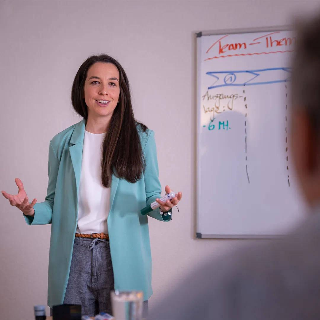 Coachingszene: Roman Wider steht neben einem Wihtboard mit er Aufschrift Team. Er lacht in Richtung der Zuhörer, die auf dem Bild nicht zu sehen sind. 