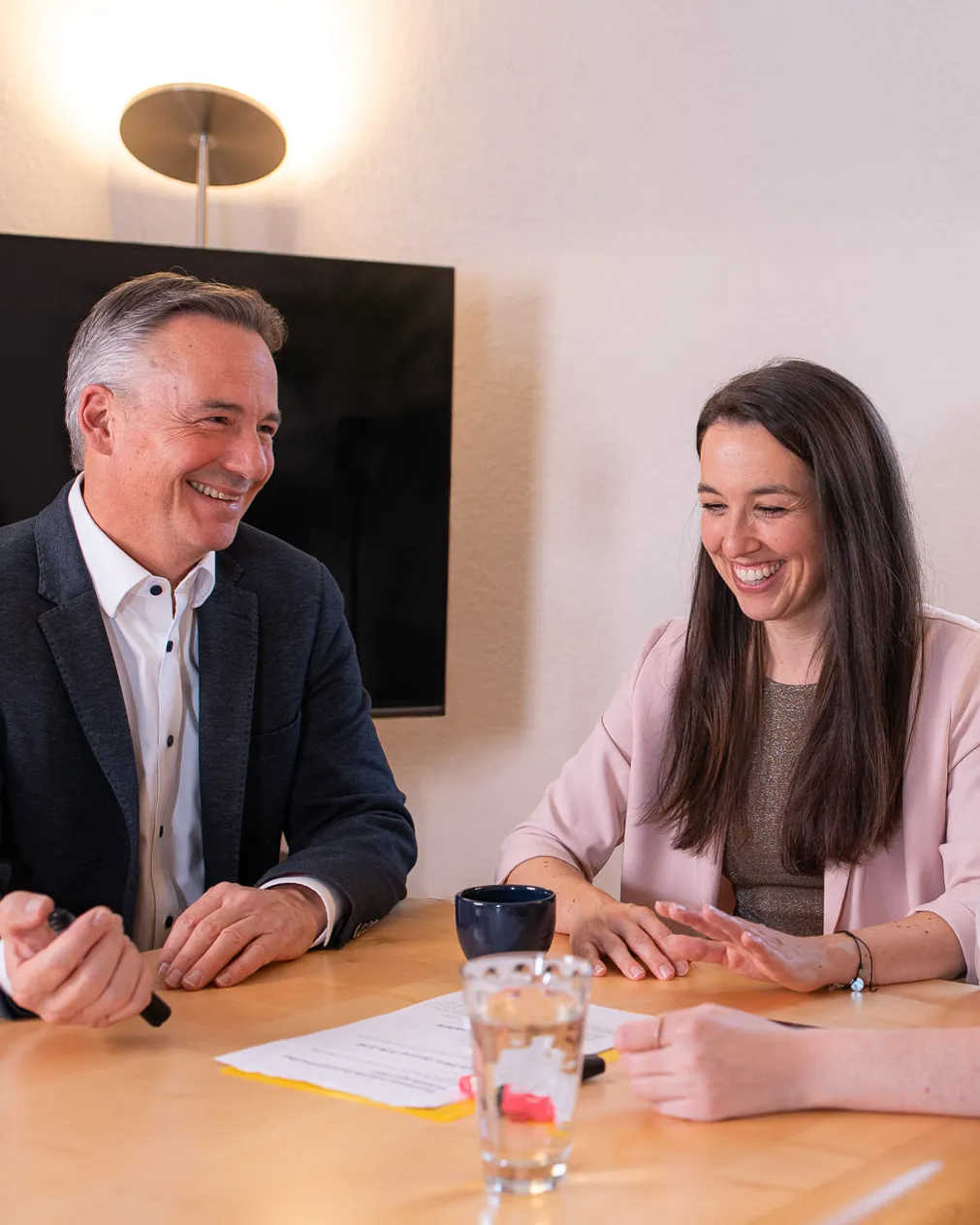 Roman und Carina sitzen an einem Tisch und besprechen etwas. Eine Hand einer dritten Person ist zu sehen.