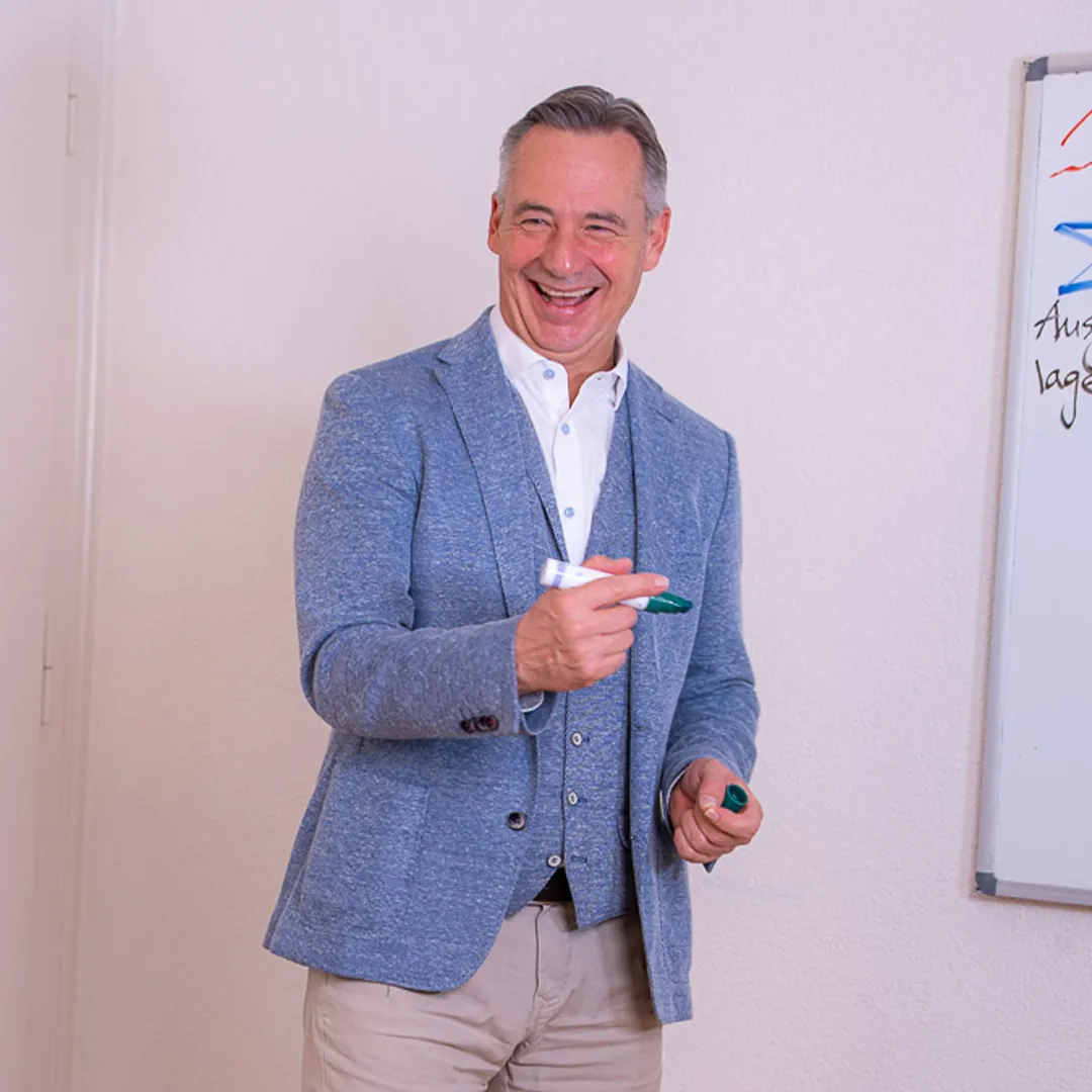 Coachingszene: Roman Wider steht neben einem Wihtboard mit er Aufschrift Team. Er lacht in Richtung der Zuhörer, die auf dem Bild nicht zu sehen sind. 