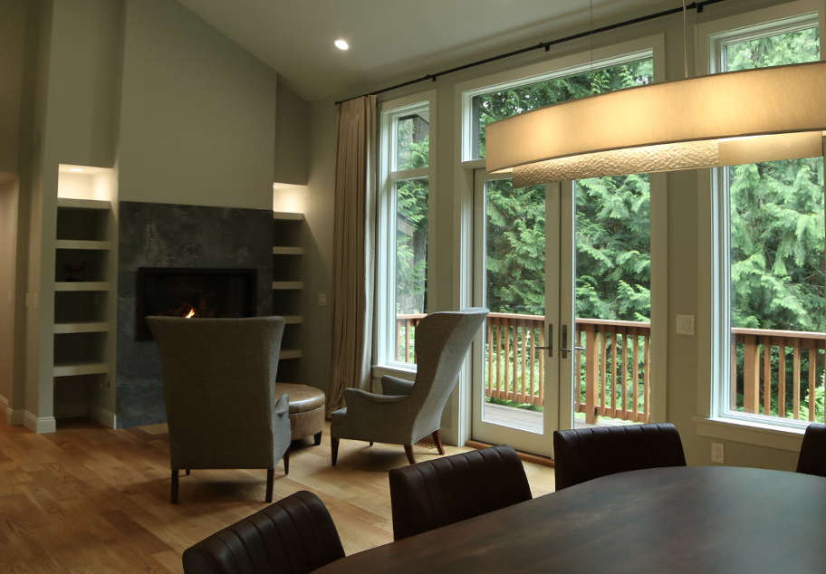 Cozy living room with two armchairs facing a lit fireplace, large windows, and a wooden deck overlooking green trees.