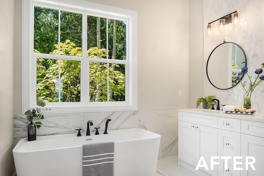 Modern bathroom with a white freestanding bathtub beneath a window showing green foliage, adjacent to a white vanity with round mirror and flower vase.