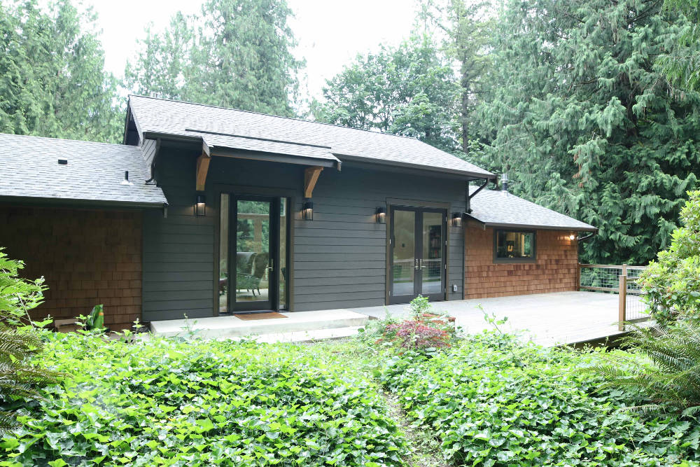 Modern house with dark siding and wooden shingles surrounded by lush green plants and tall trees.