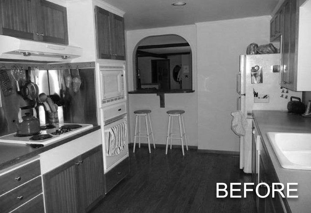 Black and white photo of a kitchen with wooden cabinets, a stove with kettle, a built-in oven and microwave, two bar stools, and a refrigerator with magnets.