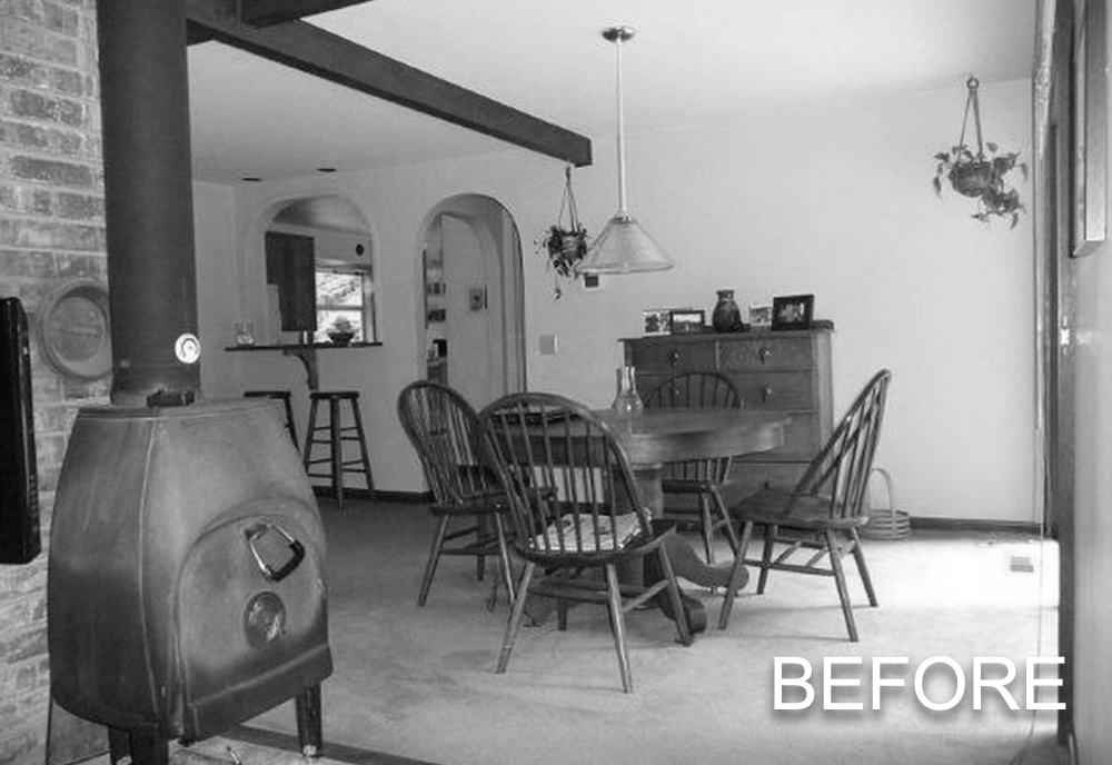 Black and white photo of a dining room with a round wooden table, four chairs, a hanging lamp, a wood stove, and a sideboard with decorations.