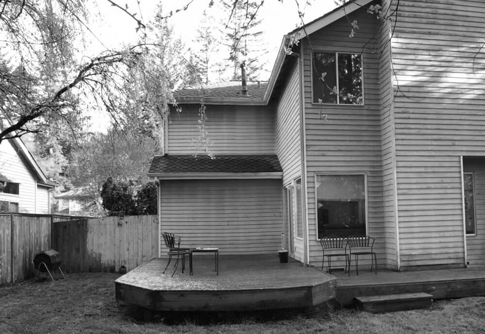 Backyard view featuring a wooden deck attached to a two-story house with outdoor chairs and a small table.