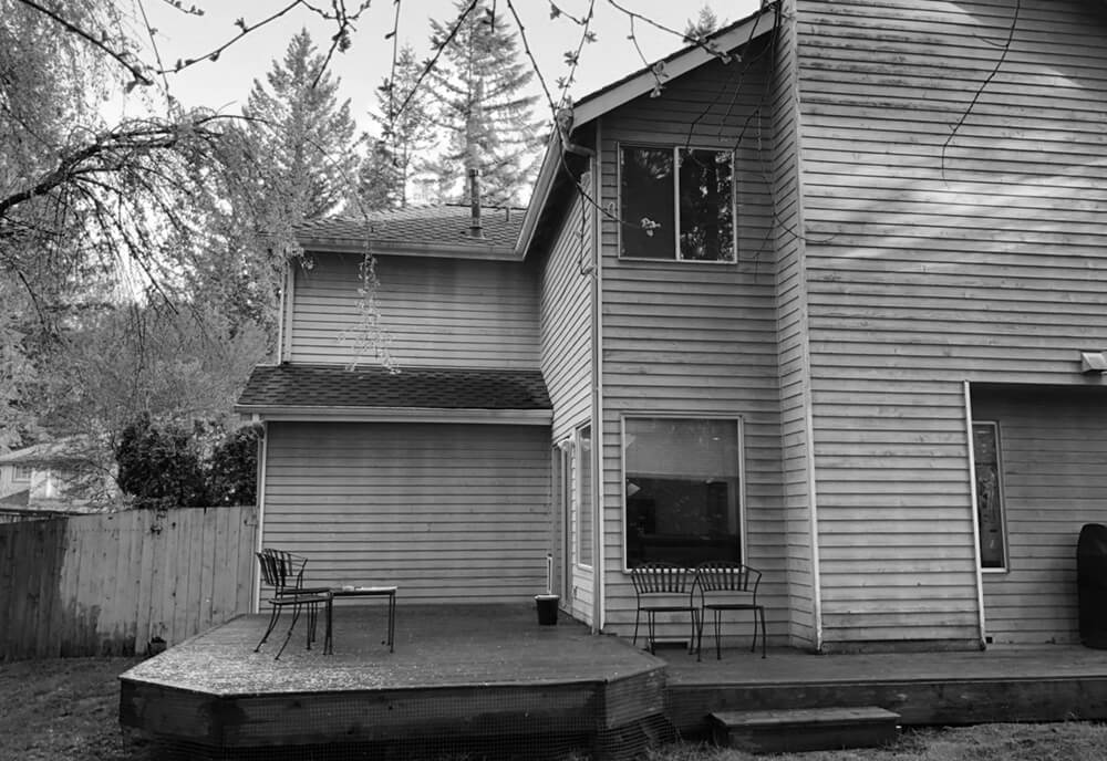Backyard of a two-story wooden house with a raised wooden deck, metal chairs, and trees in the background.