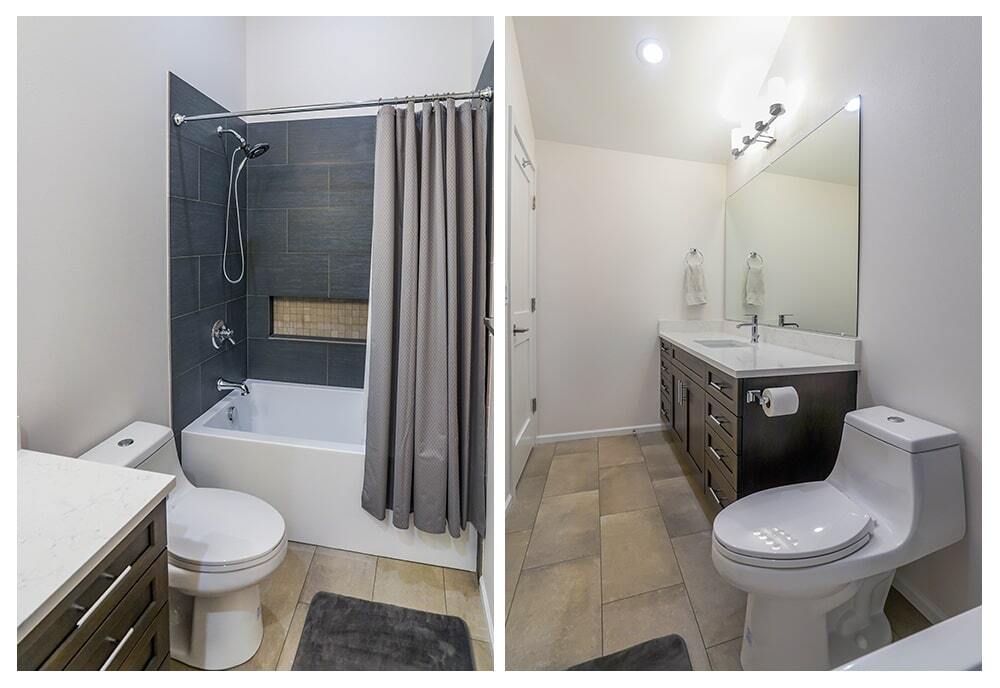 Modern bathroom with white toilet, dark wood vanity with double sinks, large mirror, and bathtub with dark gray tile surround and gray shower curtain.