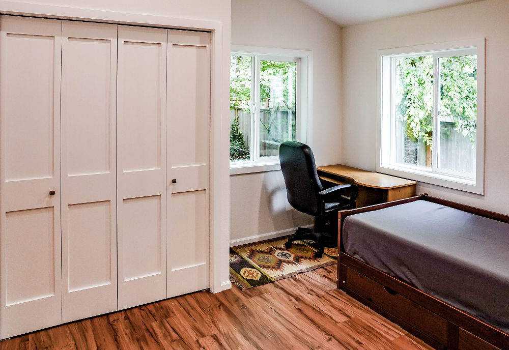 Minimalist bedroom with wooden floor, closed white closet doors, a dark wood bed with gray bedding, a corner wooden desk, black office chair, two large windows showing green foliage outside.