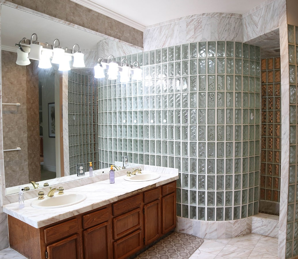 Bathroom with marble countertop, double sinks, wooden cabinets, large mirror, and curved glass block shower enclosure.